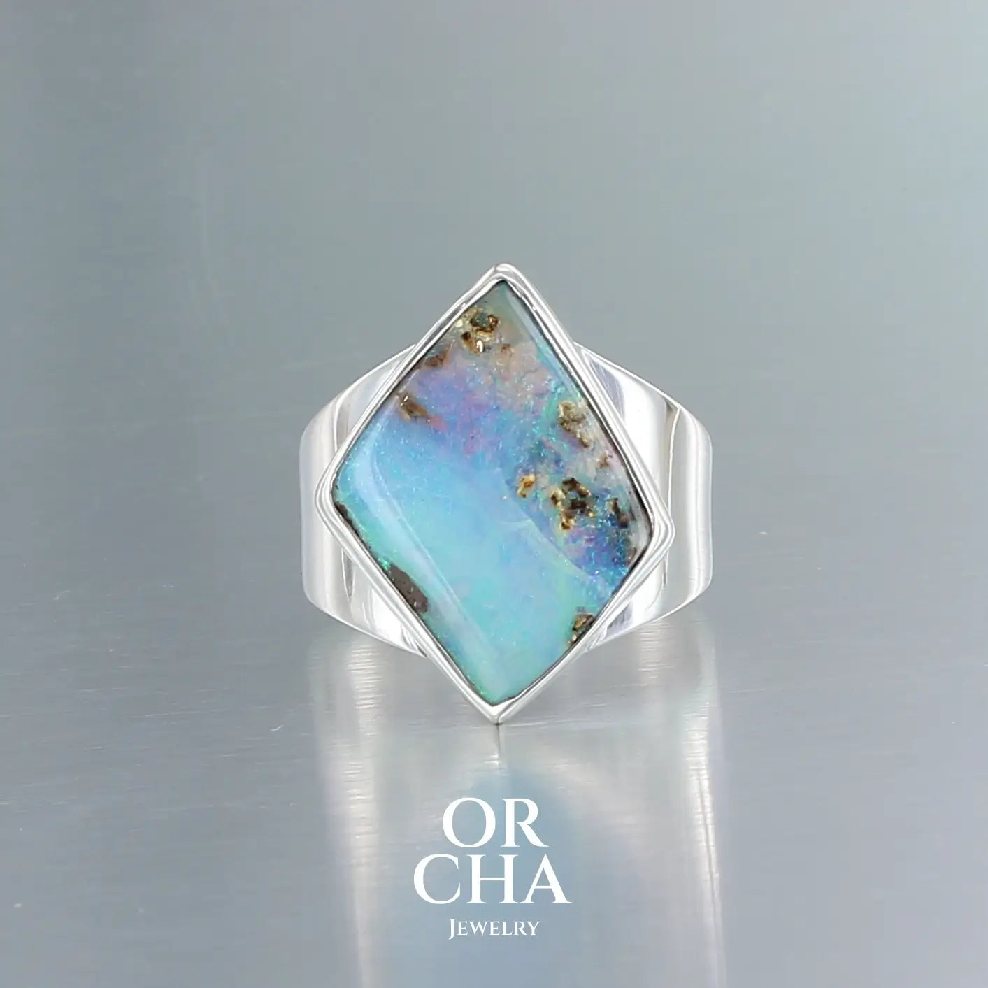 Bague en argent massif 925 sertie d'une opale boulder australienne de forme diamant asymétrique. La pierre présente une magnifique iridescence bleu-vert dominante avec des reflets violacés et quelques inclusions dorées naturelles. La monture simple et élégante en argent poli encadre la pierre, mettant en valeur ses couleurs changeantes. Bague réalisée en taille 56. Bijoux unique, éthique et durable présenté dans un coffret de notre marque, prêt à offrir. Livraison offerte, certificat d'authenticité, service
