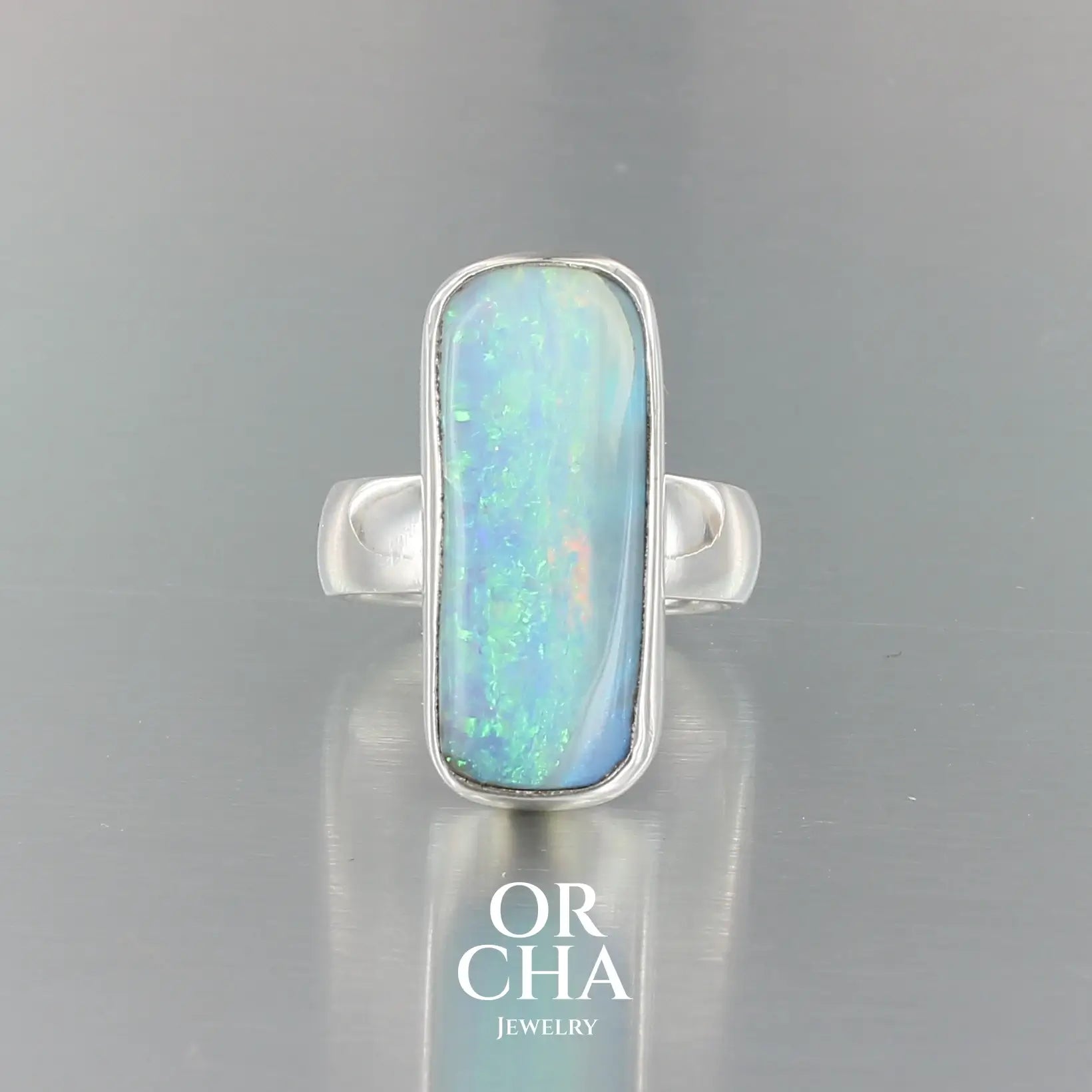 Bague en argent massif, avec une grande opale boulder rectangulaire sertie en cabochon. La pierre présente des reflets irisés bleu-vert intenses, ponctués de touches orangées. Le métal est lisse et poli, avec un anneau droit, épais et solide. Bague réalisée en taille 60. Bijoux unique, éthique et durable présenté dans un coffret de notre marque, prêt à offrir. Livraison offerte, certificat d'authenticité, service client.