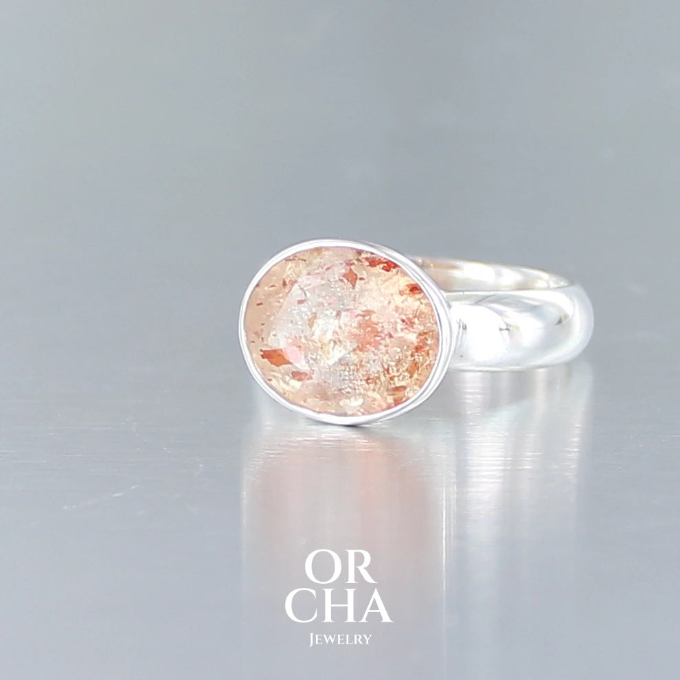 Bague cocktail pour femme en argent massif sertie d'une Pierre de soleil. Pierre ovale facettée de de couleur orange clair avec des inclusions scintillantes d'hématites. Pierre naturelle. Bijoux unique, réalisé en taille 60, éthique et durable présenté dans un coffret de notre marque, prêt à offrir. Livraison offerte, certificat d'authenticité, service client.