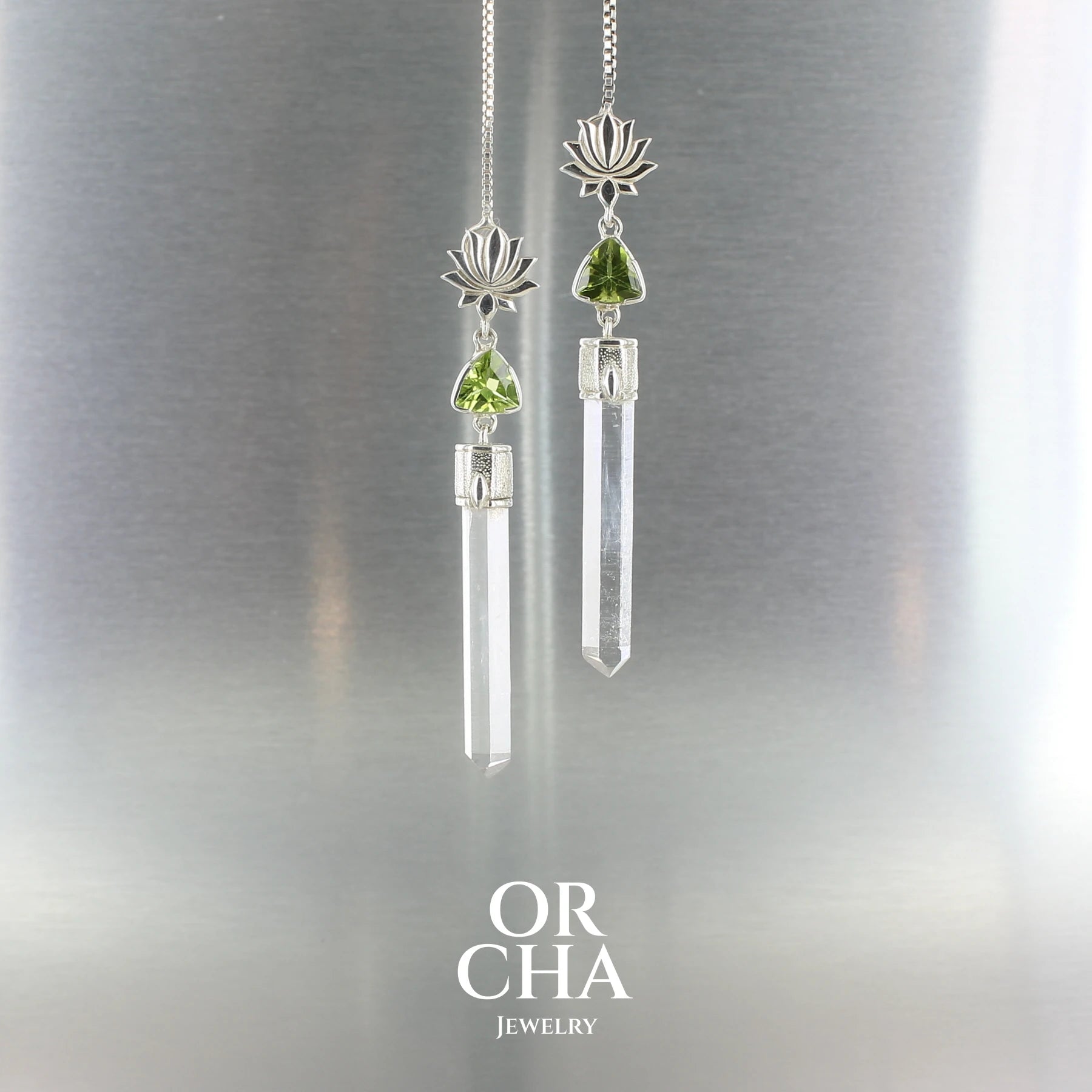 Paire de boucles d’oreilles pendantes en argent massif, ornées d’un motif de lotus stylisé sur le lobe. Sous le lotus, une petite gemme taillée de couleur vert — un péridot — est sertie de manière délicate. Elle est suivie d’un cristal de roche naturel, long et fin, transparent, non taillé, en forme de pointe verticale. L’ensemble forme une silhouette élancée, à la fois pure et élégante.
