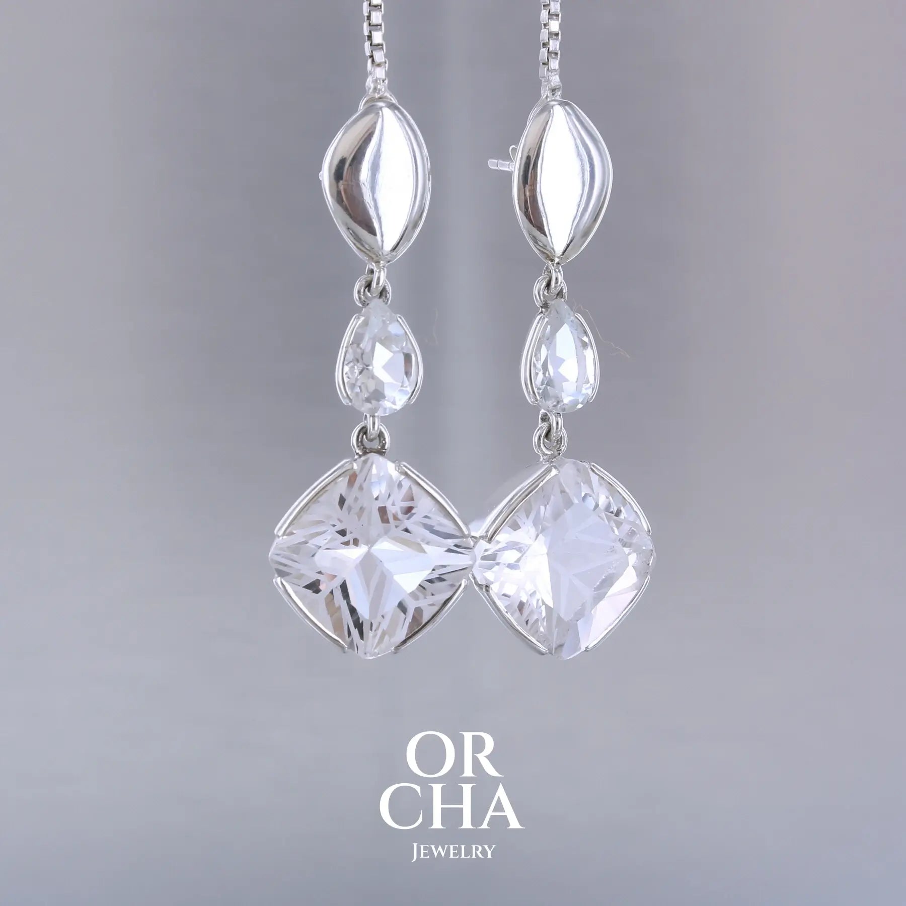 Boucles d'oreilles pendantes pour femme en argent massif serties de Cristals de roche gravés d'étoiles et de topazes blanches. Pierres facettée de forme carrée et goutte de qualité transparente, propre à l'oeil. Pierre naturelle incolore et blanche sur les parties gravées. Boucles de 3,7 cm de long. Bijoux unique, éthique et durable présenté dans un coffret de notre marque, prêt à offrir. Livraison offerte, certificat d'authenticité, service client.