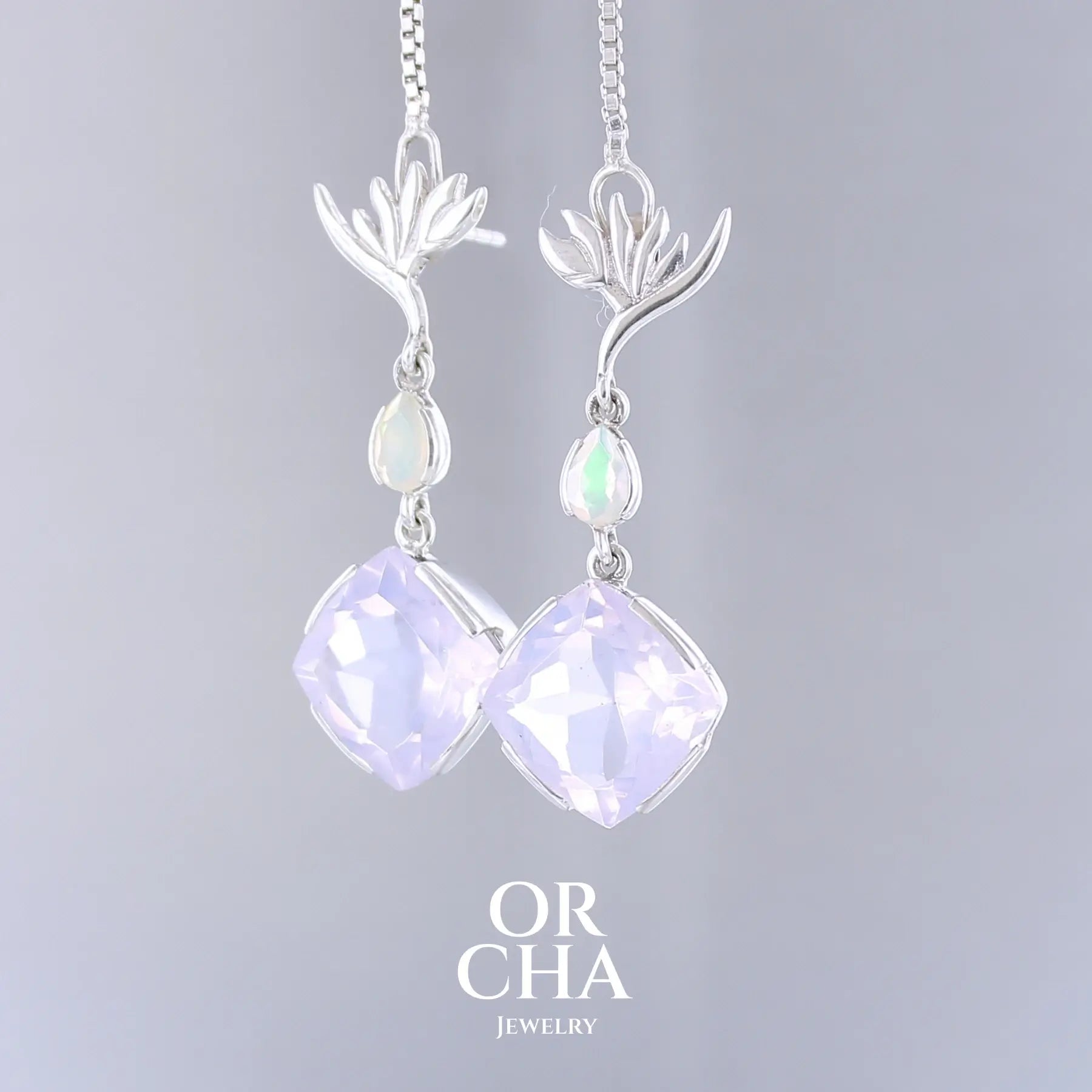 Boucles d'oreilles Strelitzia pour femme en argent massif sertie de Quartz lavandes et d'opales d'Éthiopie. Pierres facettées de forme ovales et de gouttes de qualités transparentes, propre à l'oeil. Pierres naturelles de couleurs violettes pastel teintées de rose avec des reflets opalescents et multicolores pour les opales. Bijoux unique, éthique et durable présenté dans un coffret de notre marque, prêt à offrir. Livraison offerte, certificat d'authenticité, service client.