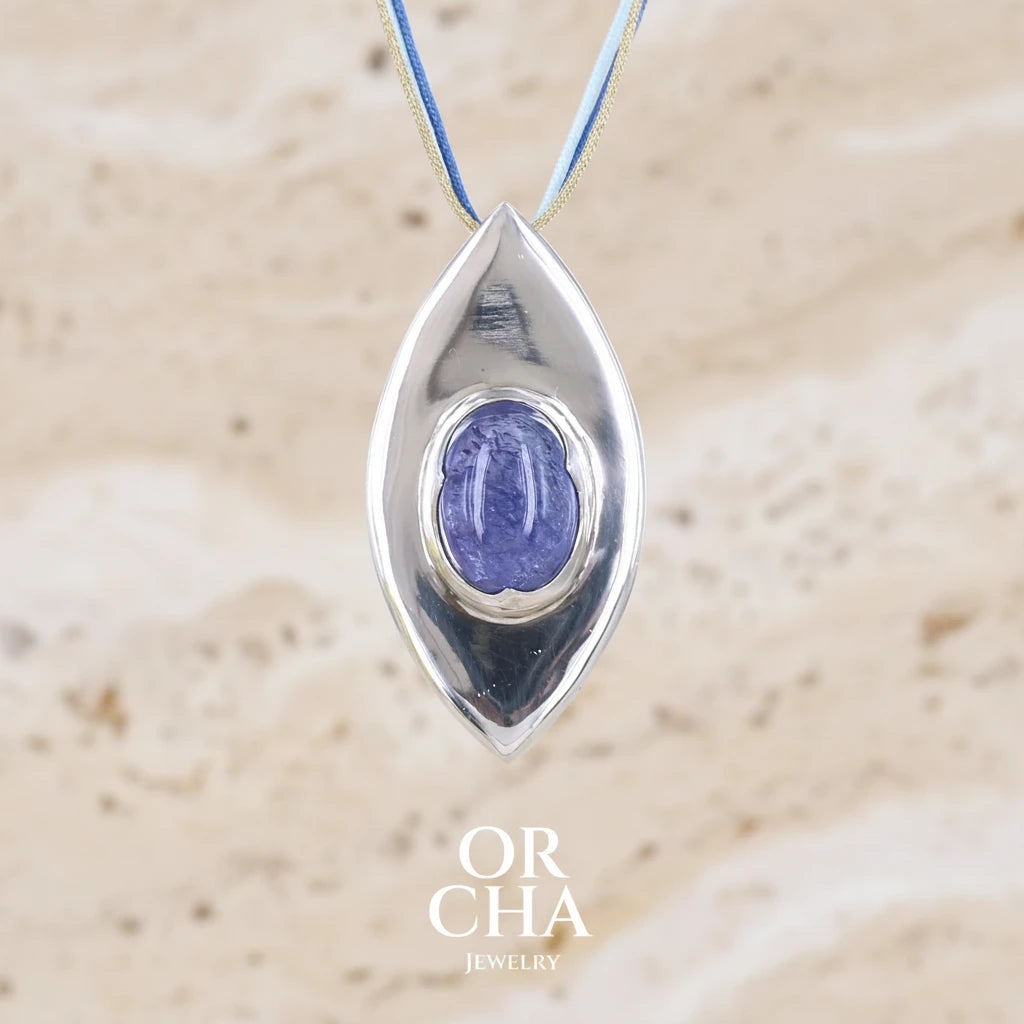 Sautoir luxe fantaisie pour femme en corde et argent massif sertie d'une Tanzanite. Pierre cabochon de forme ovale de qualité transparente à inclusions. Pierre  de couleur bleu lavande. Pendentif monté sur 3 cordes de couleur bleu nuit, bleue clair et or d'une longueur total de 41 cm porté. Bijoux unique, éthique et durable présenté dans un coffret de notre marque, prêt à offrir. Livraison offerte, certificat d'authenticité, service client.