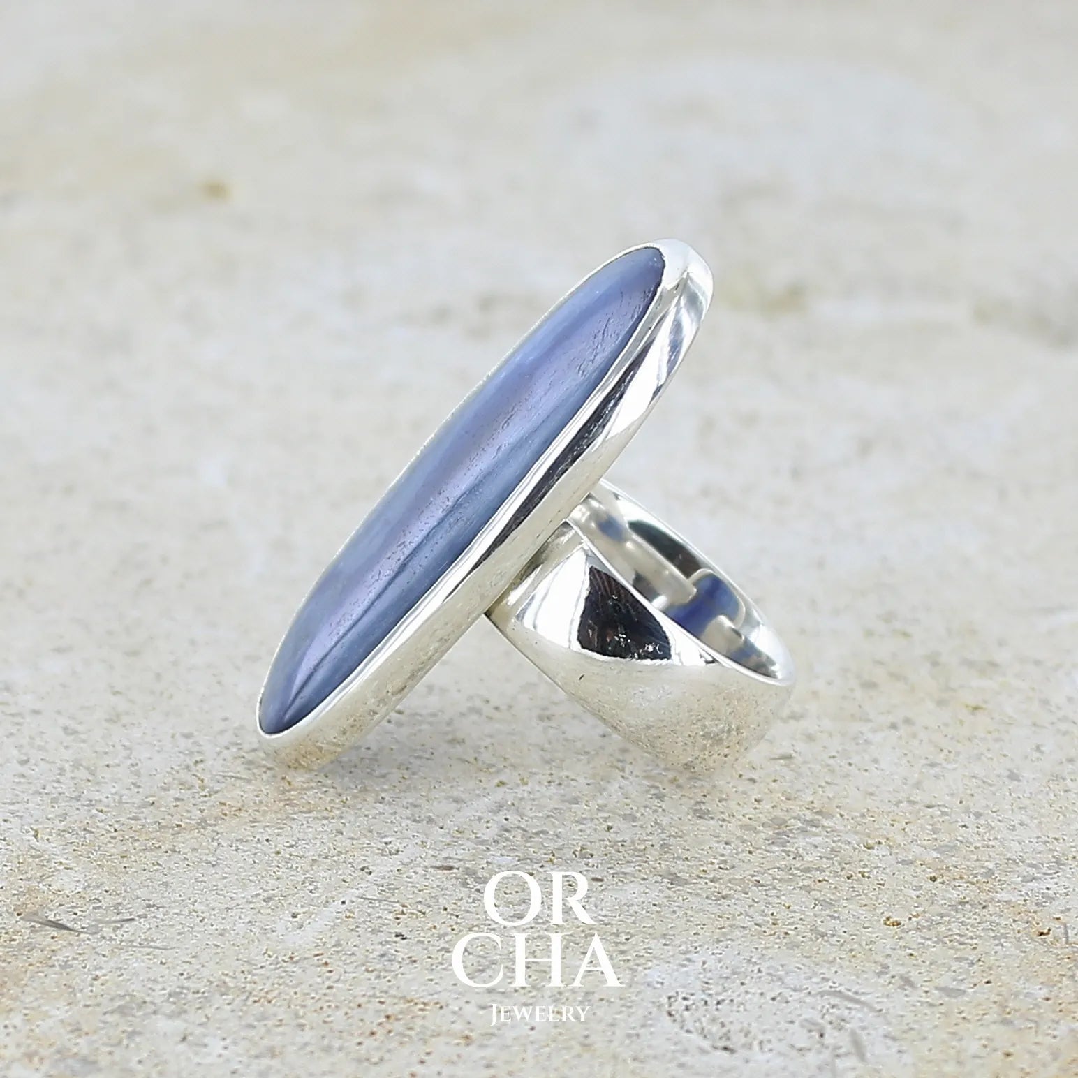 Détail du sertissage d'une bague pour femme en argent massif sertie d'un long cabochon ovale de cyanite bicolore de couleur bleu jean nacré. Pierre translucide à inclusions. Bijou unique réalisée en taille 54 par Orchajewelry
