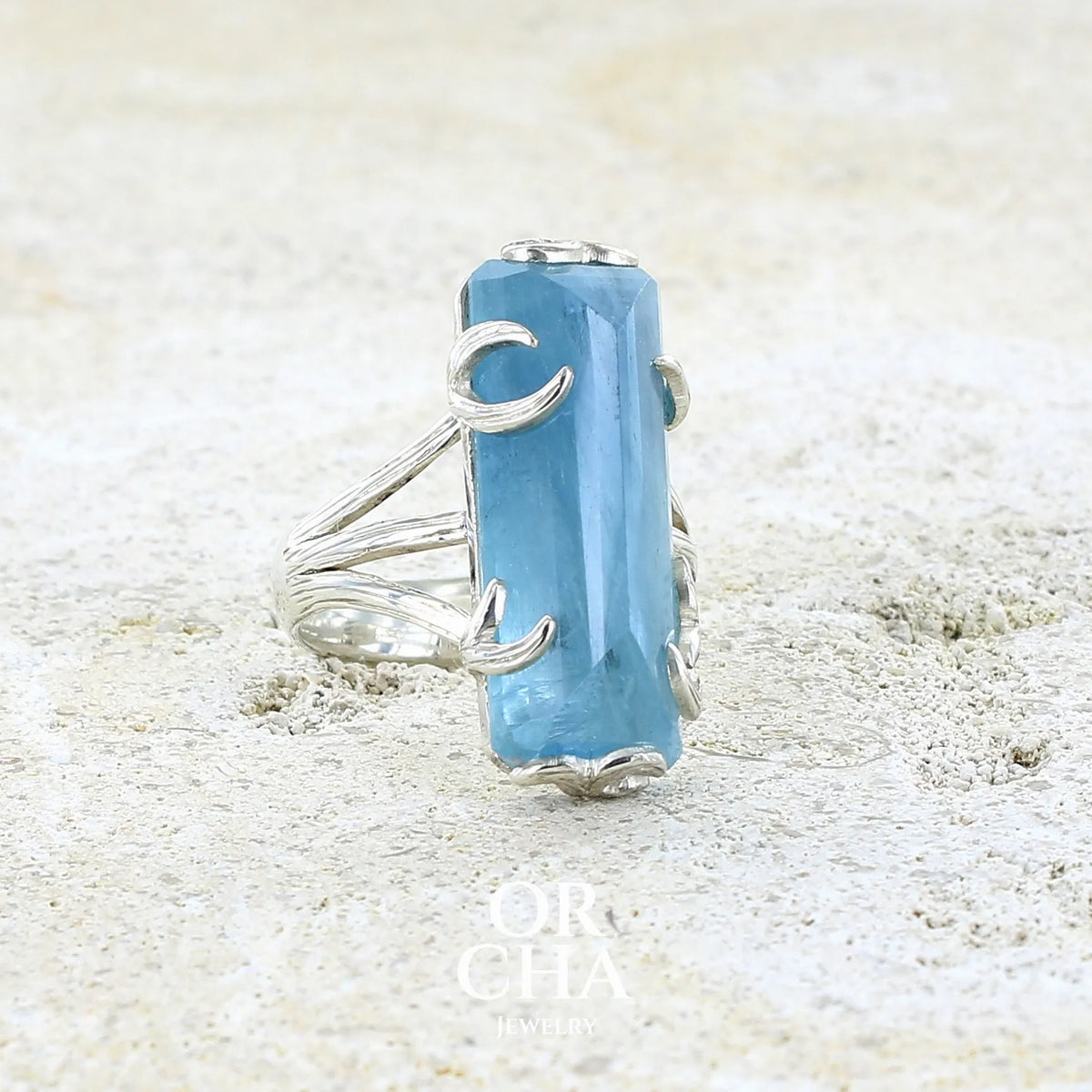 Bague cocktail pour femme en argent massif sertie d'une aigue marine taillée de 15 carats de couleur bleue gris trouble, pierre transparente avec inclusions. Pierre naturelle facettée de forme rectangulaire. Bijoux unique réalisée en taille 54,5 par Orchajewelry 