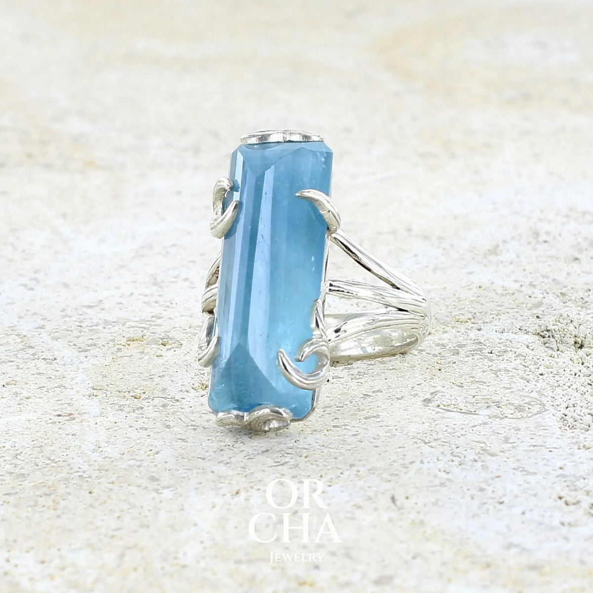 Bague cocktail pour femme en argent massif sertie d'une aigue marine taillée de 15 carats de couleur bleue gris trouble, pierre transparente avec inclusions. Pierre naturelle facettée de forme rectangulaire. Bijoux unique réalisée en taille 54,5 par Orchajewelry 