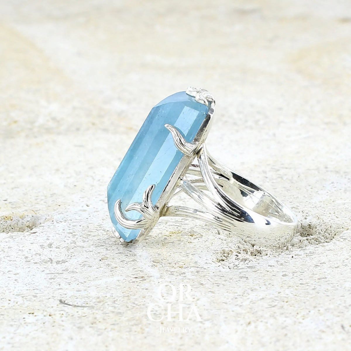 Bague cocktail pour femme en argent massif sertie d'une aigue marine taillée de 15 carats de couleur bleue gris trouble, pierre transparente avec inclusions. Pierre naturelle facettée de forme rectangulaire. Bijoux unique réalisée en taille 54,5 par Orchajewelry 