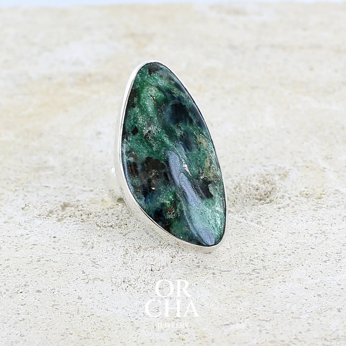 Bague pour femme en argent massif sertie d'un cabochon de pierre verte scintillante à inclusions bleues et noires. Pierre de qualité opaque et de forme libre. Bijou unique réalisée en taille 56 par Orchajewelry