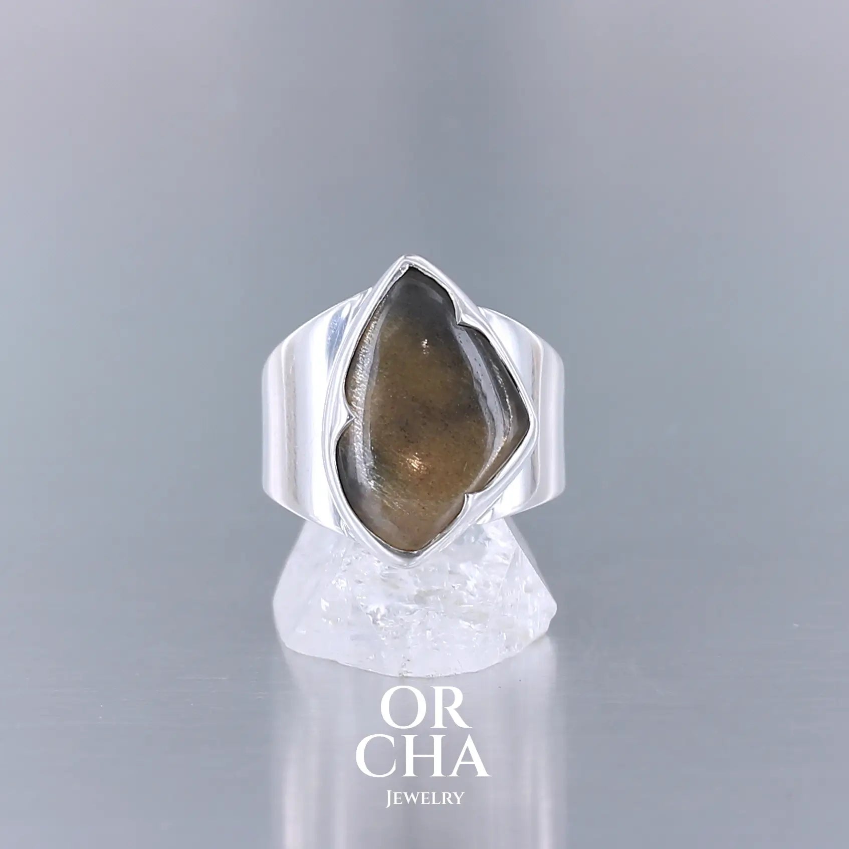 Bague en argent massif 925 sertie d'une pierre de soleil noire en cabochon, taillée en forme libre évoquant une goutte stylisée. La gemme sombre présente des reflets métalliques cuivrés caractéristiques, mis en valeur par un serti clos à pointe et un anneau large poli miroir. Design artisanal contemporain mettant en lumière l’éclat unique de la pierre naturelle. Bijoux, éthique et durable présenté dans un coffret de notre marque, prêt à offrir. Livraison offerte, certificat d'authenticité, service client.