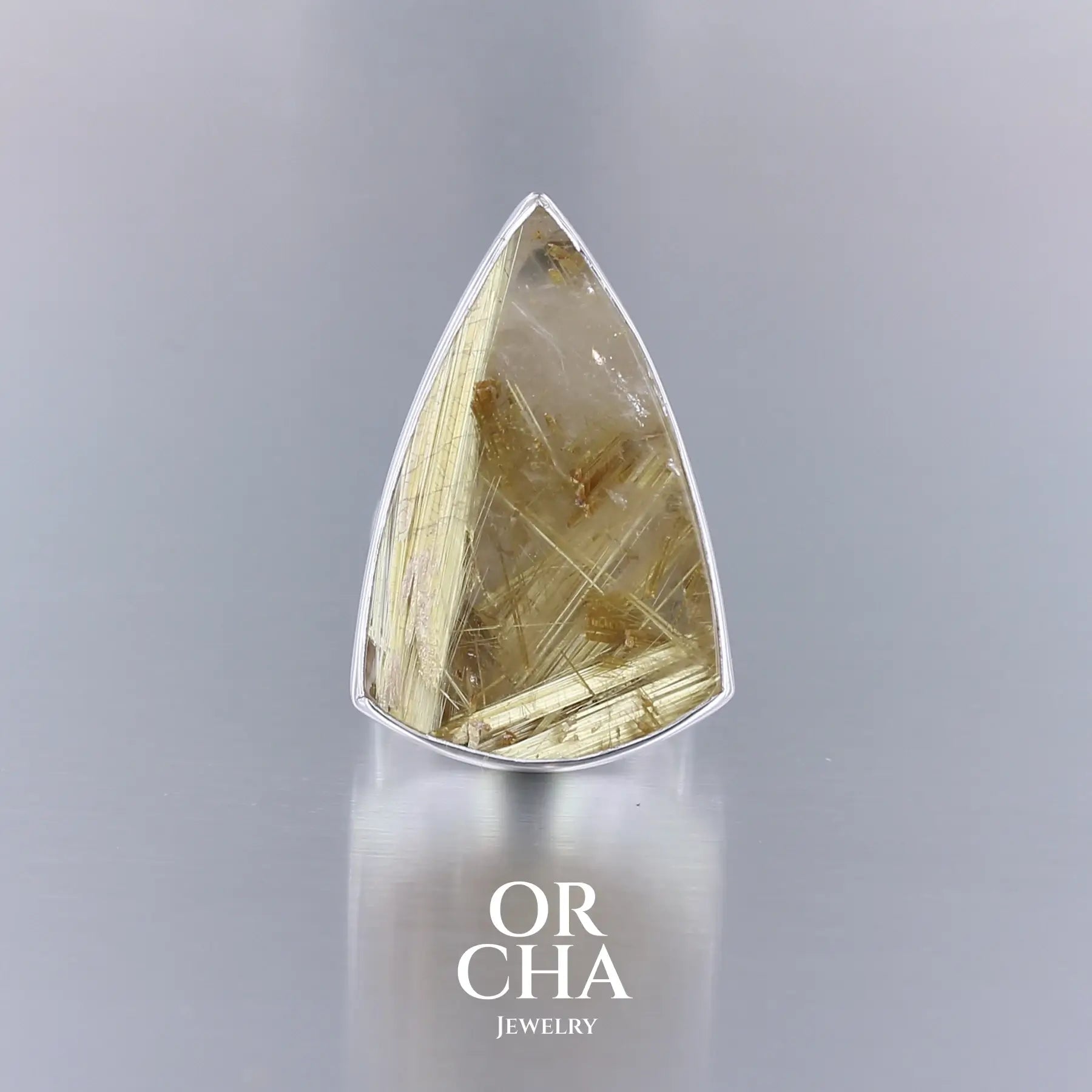 Grande bague en argent massif ornée d’un quartz rutile doré taillé en forme de bouclier allongé, aux inclusions fibreuses dorées bien visibles, formant un motif graphique complexe sous la surface brillante de la pierre. Le serti lisse suit fidèlement les contours de la gemme, mettant en valeur sa transparence et ses reflets dorés.  Bijoux unique, éthique et durable présenté dans un coffret de notre marque, prêt à offrir. Livraison offerte, certificat d'authenticité, service client.
