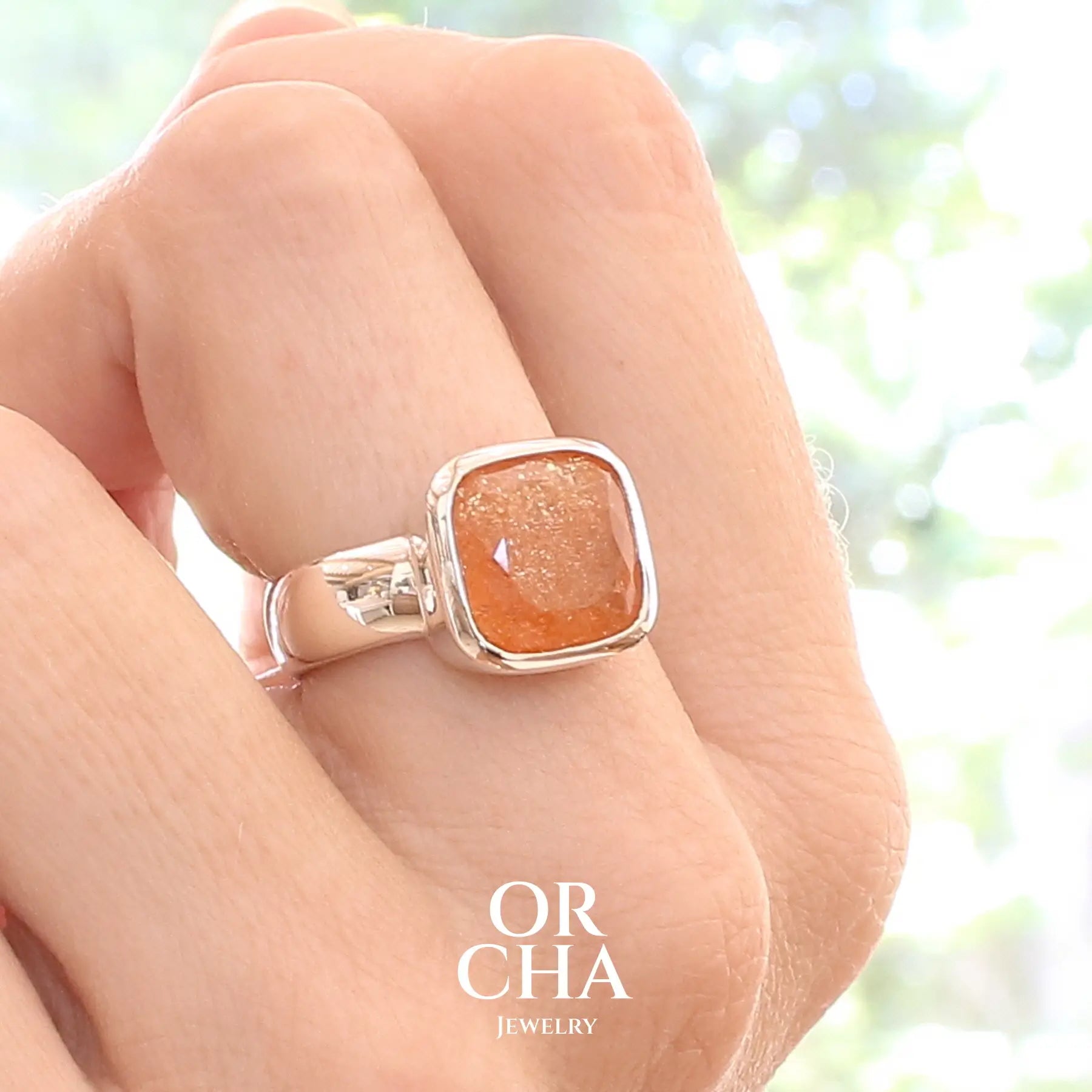 photo sur modèle d'une bague cocktail pour femme en argent massif sertie d'une pierre de soleil oranges de très belle qualité, pierre transparente avec inclusions scintillantes d'hématites. Pierre naturelle facettée de forme carrée. Tailles 56. Bijoux unique, éthique et durable présenté dans un coffret de notre marque, prêt à offrir. Livraison offerte, certificat d'authenticité, service client.