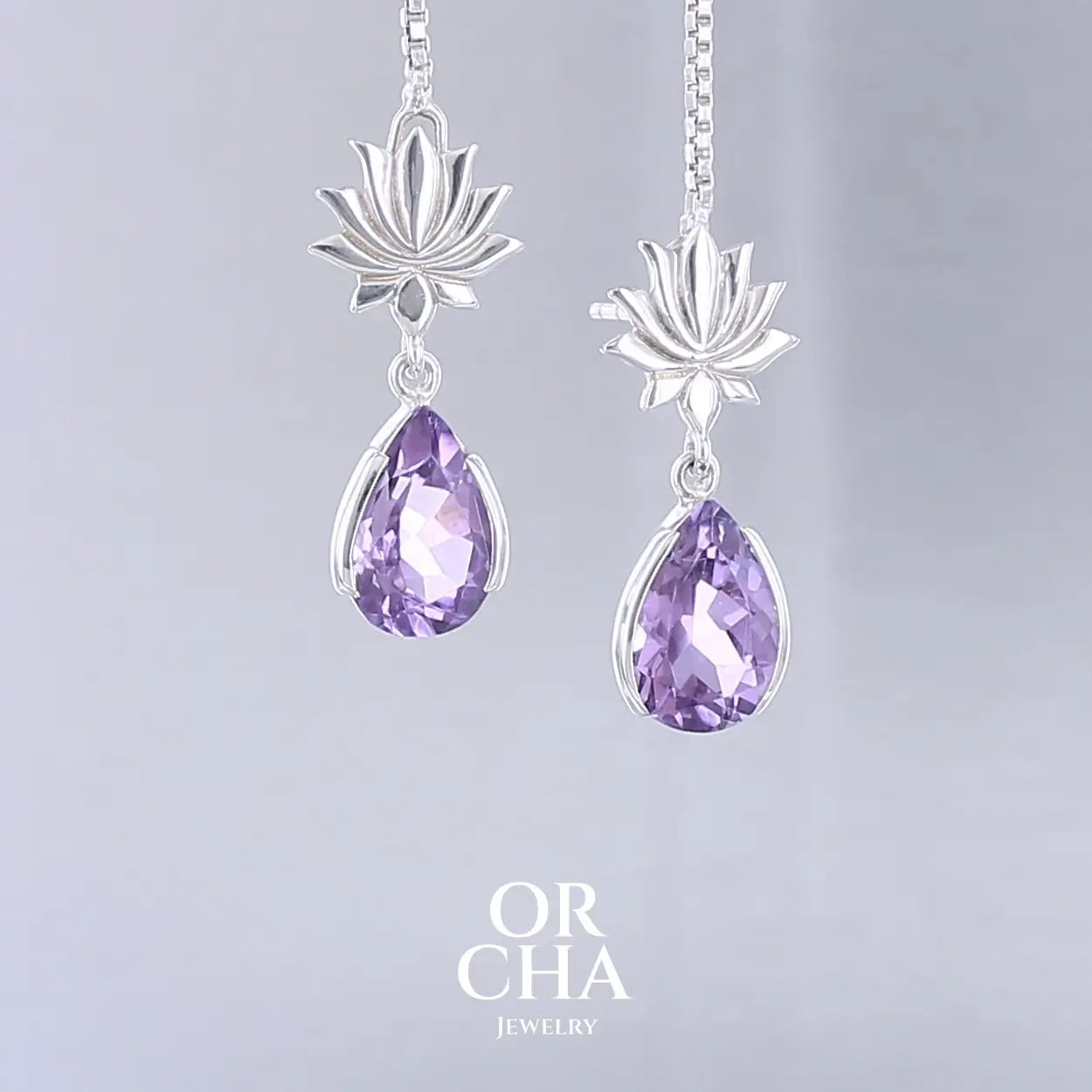 Boucles d'oreilles Lotus pour femme en argent massif serties d'Améthystes. Pierres facettées en forme de gouttes de qualités transparentes, propre à l'oeil. Pierres naturelles de couleurs violettes moyen. Bijoux unique, éthique et durable présenté dans un coffret de notre marque, prêt à offrir. Livraison offerte, certificat d'authenticité, service client.