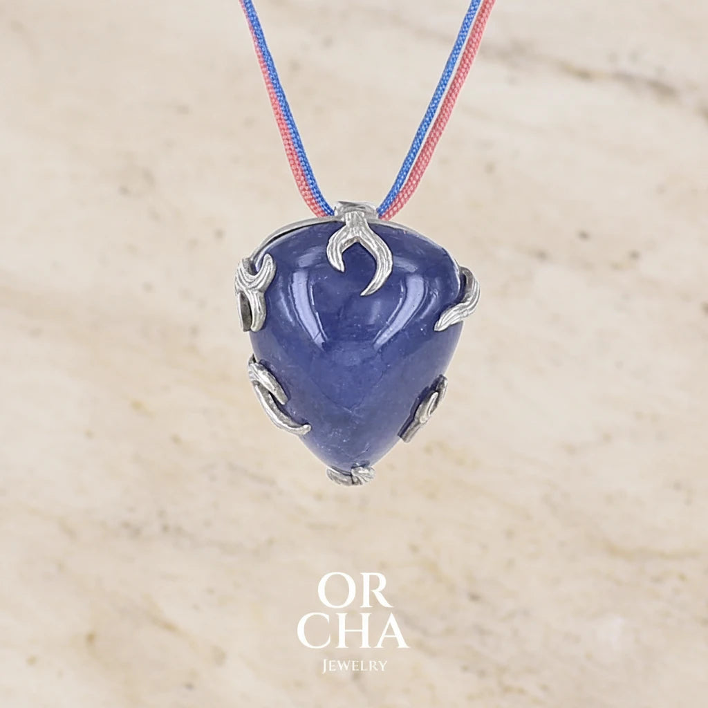 Sautoir luxe fantaisie pour femme en corde et argent massif sertie d'une Tanzanite. Pierre cabochon en forme de goutte de qualité opaque à inclusions. Pierre  de couleur bleu foncé. Pendentif monté sur 2 cordes de couleur bleu nuit, et corail d'une longueur total de 40 cm porté. Bijoux unique, éthique et durable présenté dans un coffret de notre marque, prêt à offrir. Livraison offerte, certificat d'authenticité, service client.