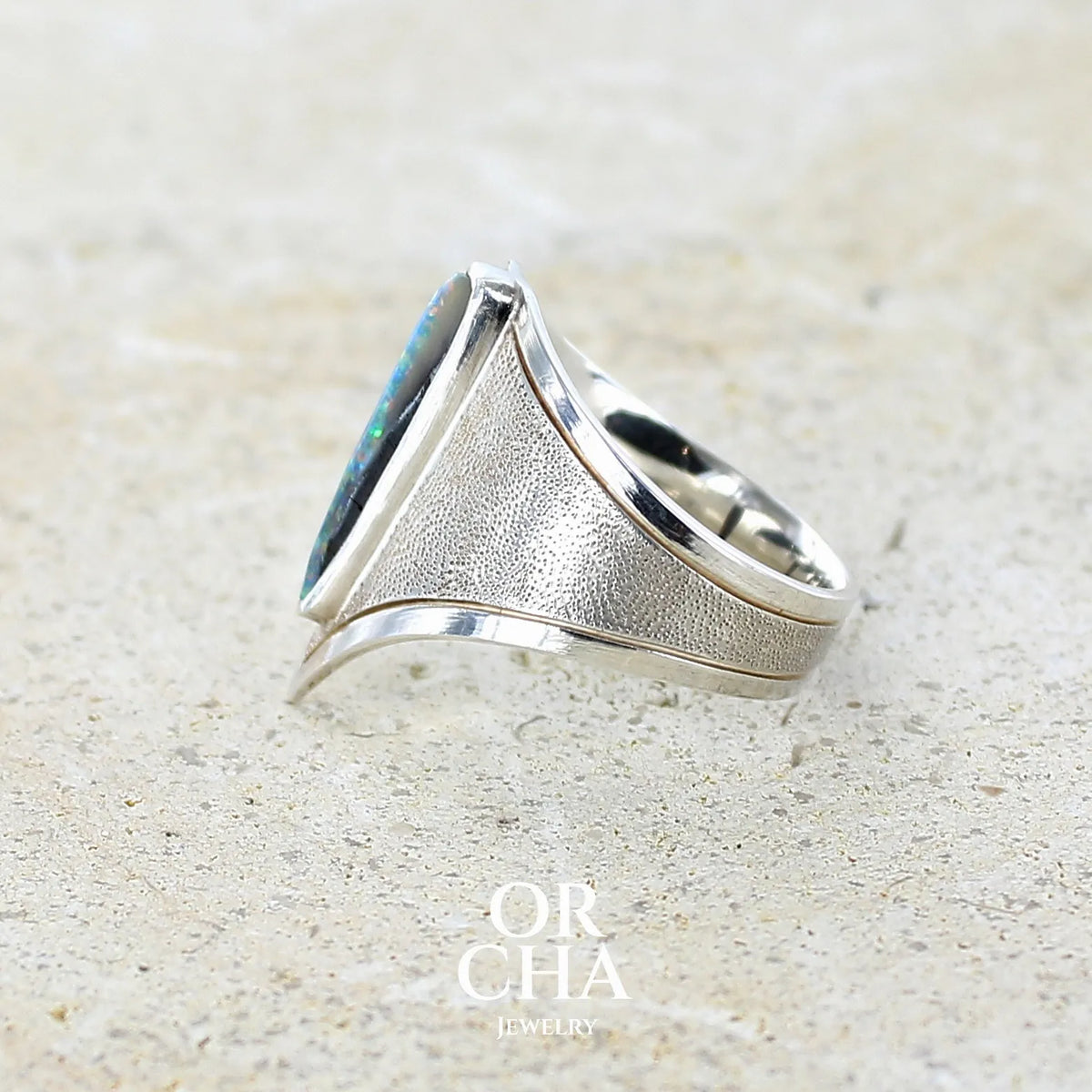 Détail d'une bague pour femme en argent massif 925 sertie d'une opale boulder aux feux multicolores bleus violet et verts. Cabochon d'opale massive naturelle taillée en forme d'aile. Pierre de qualité opaque. Bijoux unique réalisé en taille 57 par Orcha Jewelry.