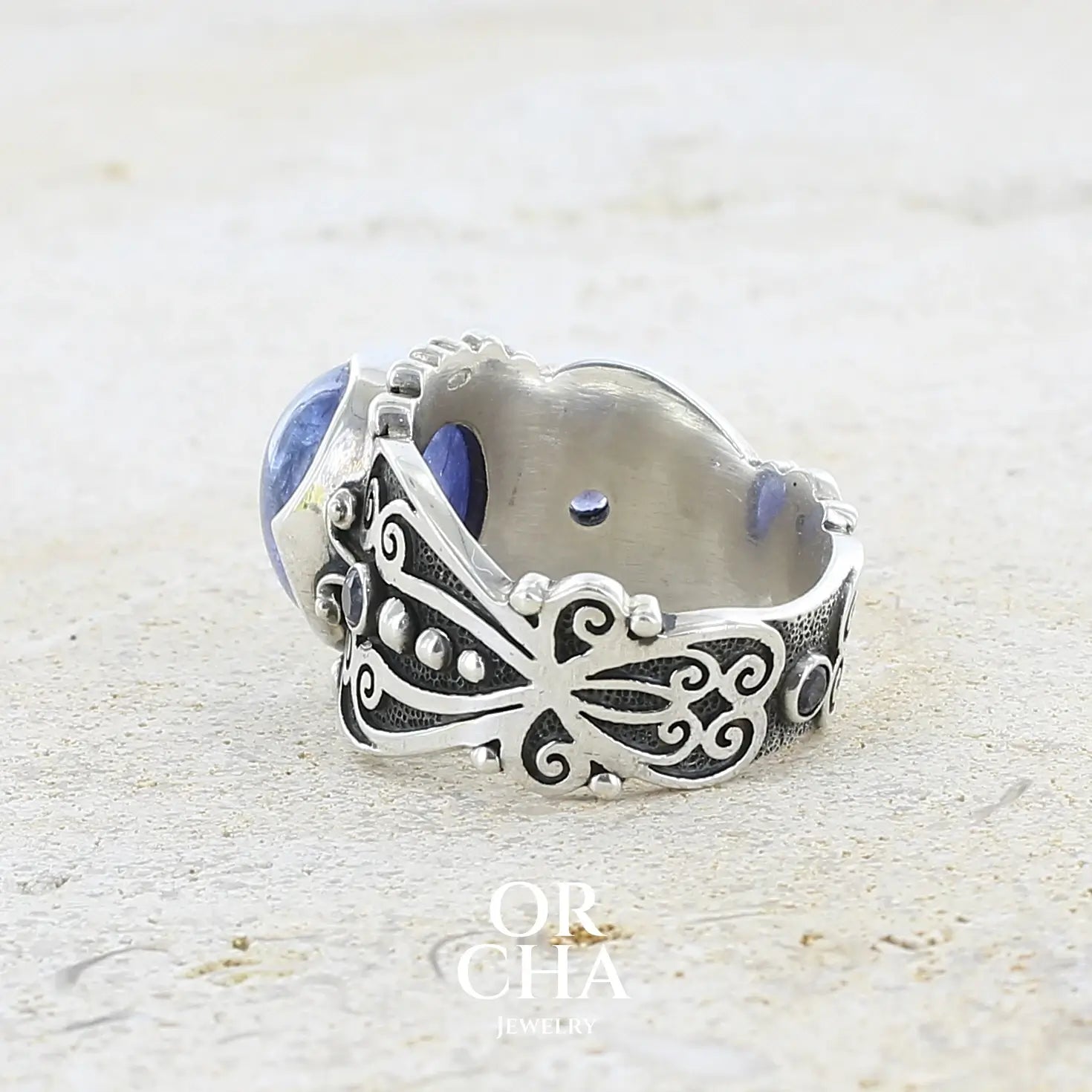 détail texture d'une bague en argent massif sculptées d'entrelacs de style celtique qui illustrent des papillons, sertie d'une Tanzanite. Pierre cabochon de forme ovale, de couleur bleu lavande foncé, de qualité transparente avec inclusions. Taille 56. Bijoux unique, éthique et durable présenté dans un coffret de notre marque, prêt à offrir. Livraison offerte, certificat d'authenticité, service client.
