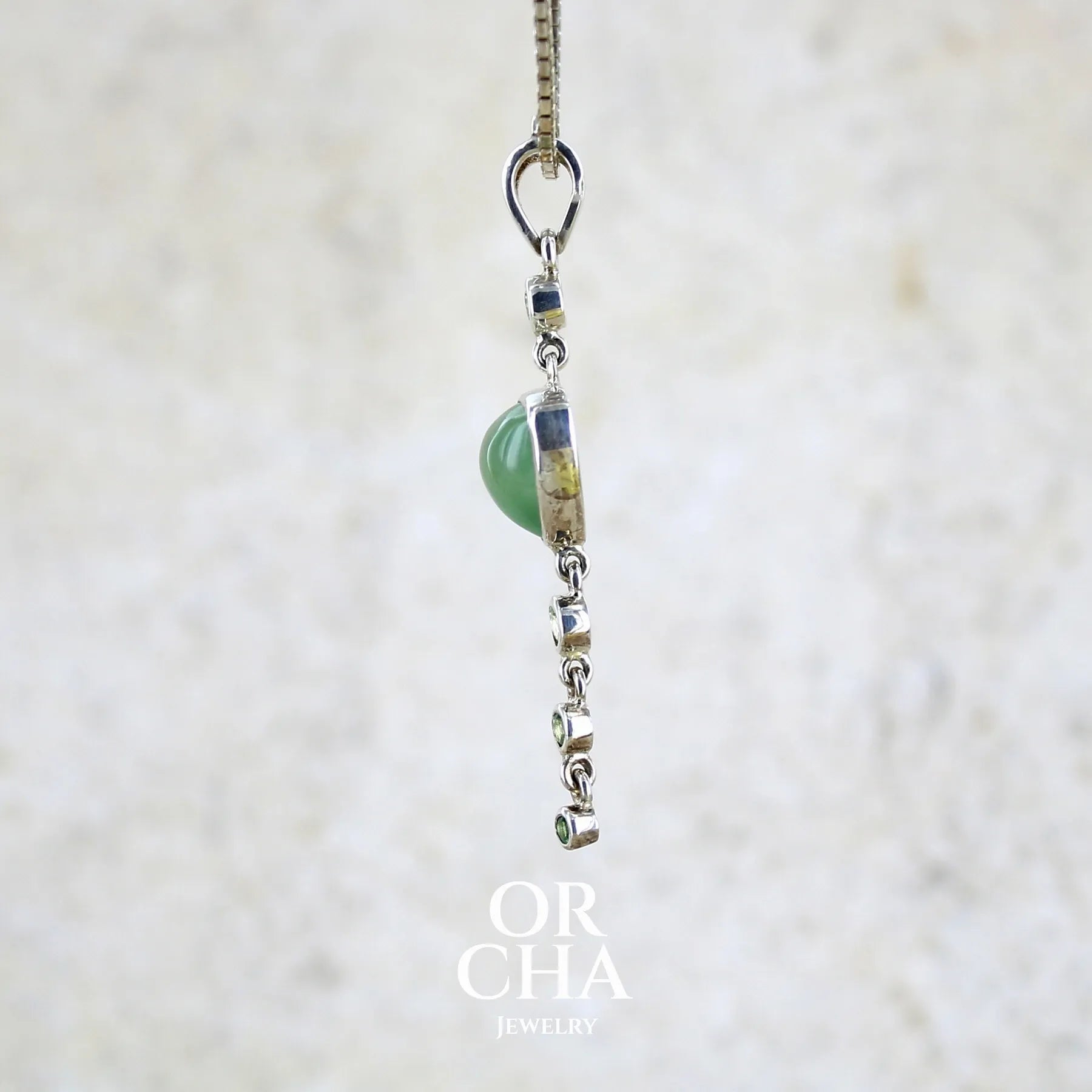 Détail du sertissage d'un pendentif pour femme en argent massif sertie d'un cabochon triangulaire de Préhnite ainsi que de petites tourmalines vertes formant une cascade colorée vert pastel lumineux. Pierre transparente à inclusions. Bijou unique réalisé par Orchajewelry