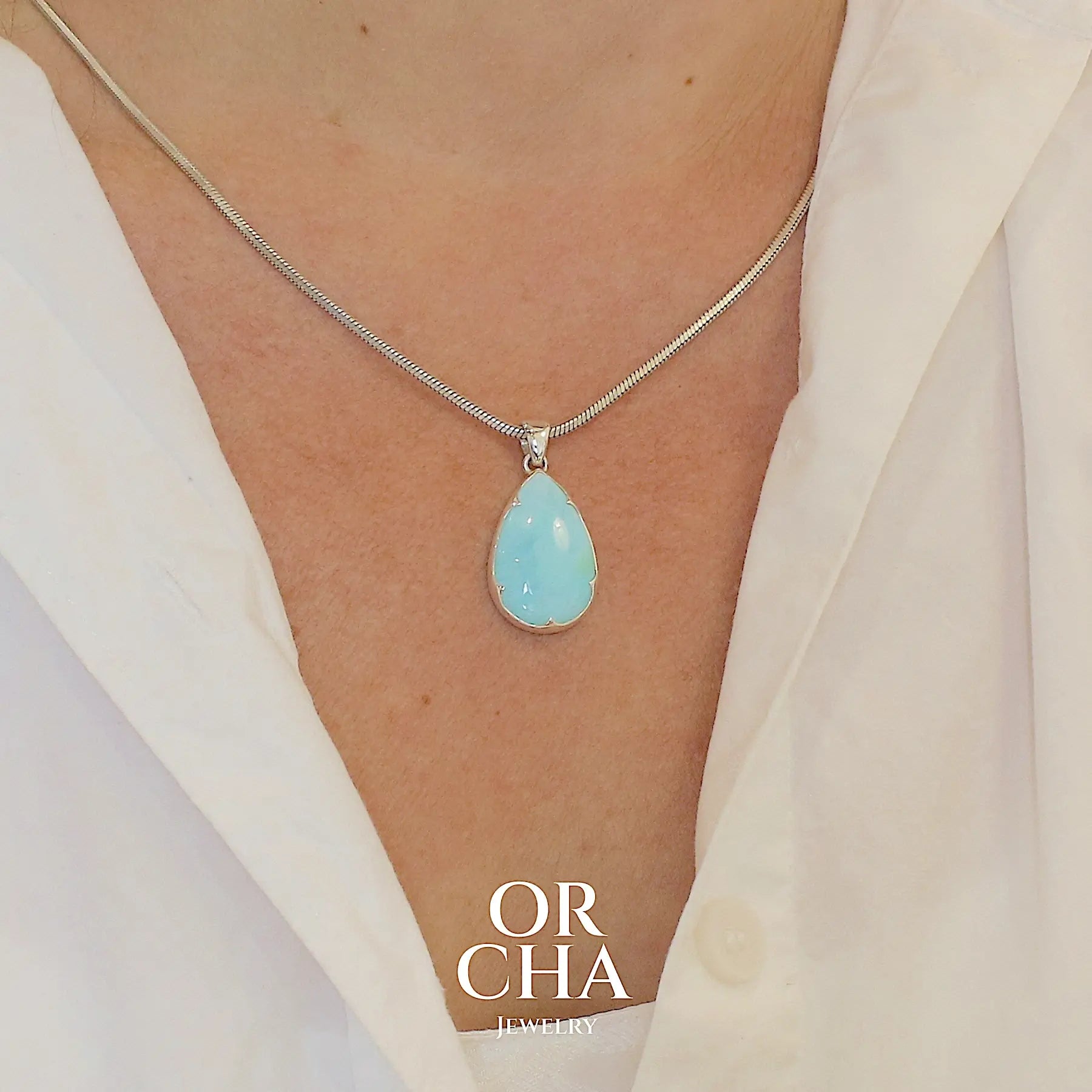photo sur modèle d'un pendentif en argent massif, sertie d'un cabochon d'opale bleue du Pérou de couleur bleu ciel clair. Pierre en forme de goutte. Pierre opaque et translucide. Bijoux unique, éthique et durable présenté dans un coffret de notre marque, prêt à offrir. Livraison offerte, certificat d'authenticité, service client.