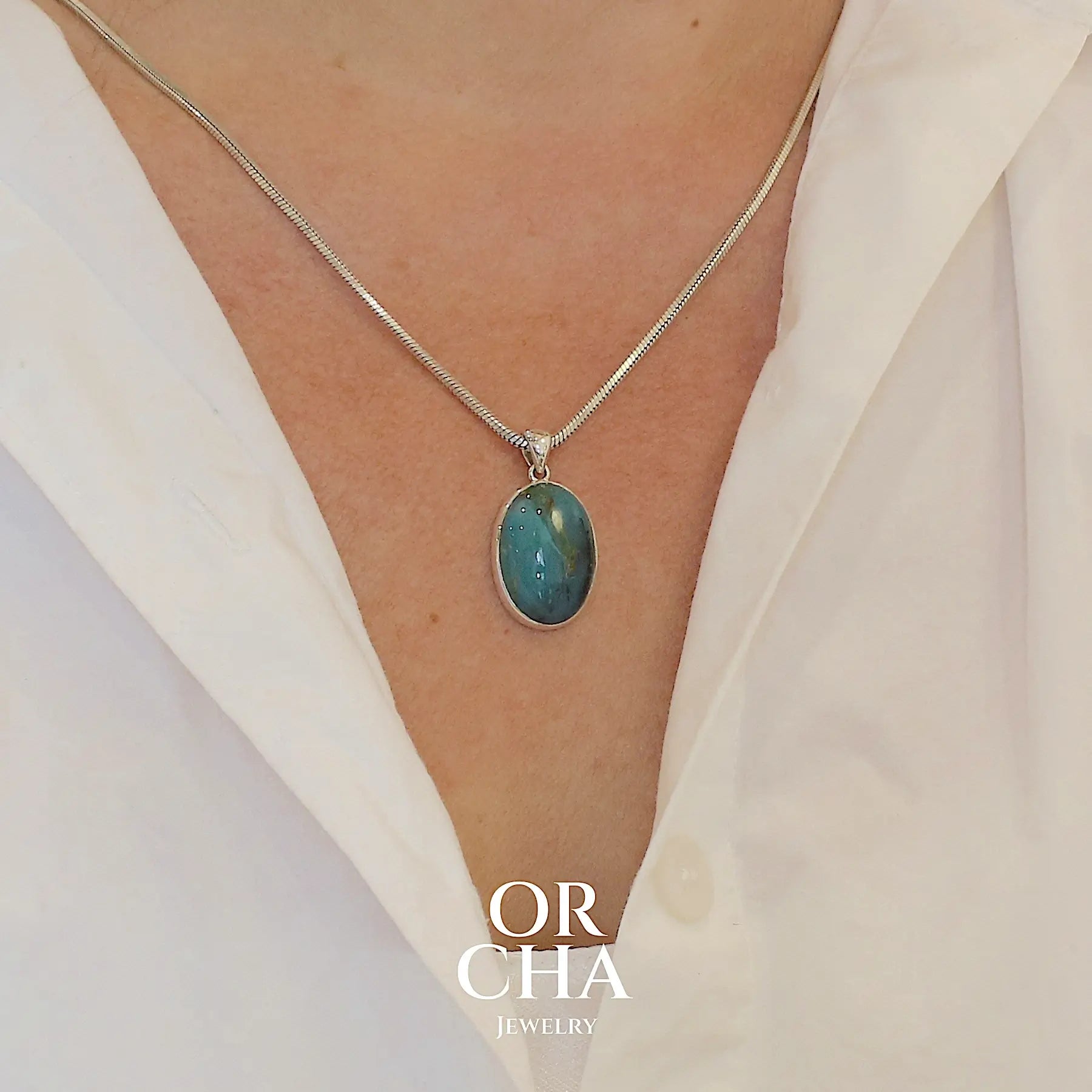 photo sur modèle d'un pendentif en argent massif, sertie d'un cabochon ovale d'opale bleue du Pérou de couleur bleu laiteux foncé à inclusions de dendrites paysages noires et de roche mère marron. Pierre opaque et translucide. Bijoux unique, éthique et durable présenté dans un coffret de notre marque, prêt à offrir. Livraison offerte, certificat d'authenticité, service client.