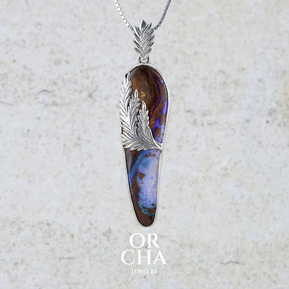 pendentif long en argent massif serti d'un cabochon d'Opale Boulder. Pierre naturelle au feux bleus verts et violets inclus dans une roche mère marron. Le sertissage et la bélière reprennent le thème de l'épi de blé. Bijou unique par Orcha jewelry