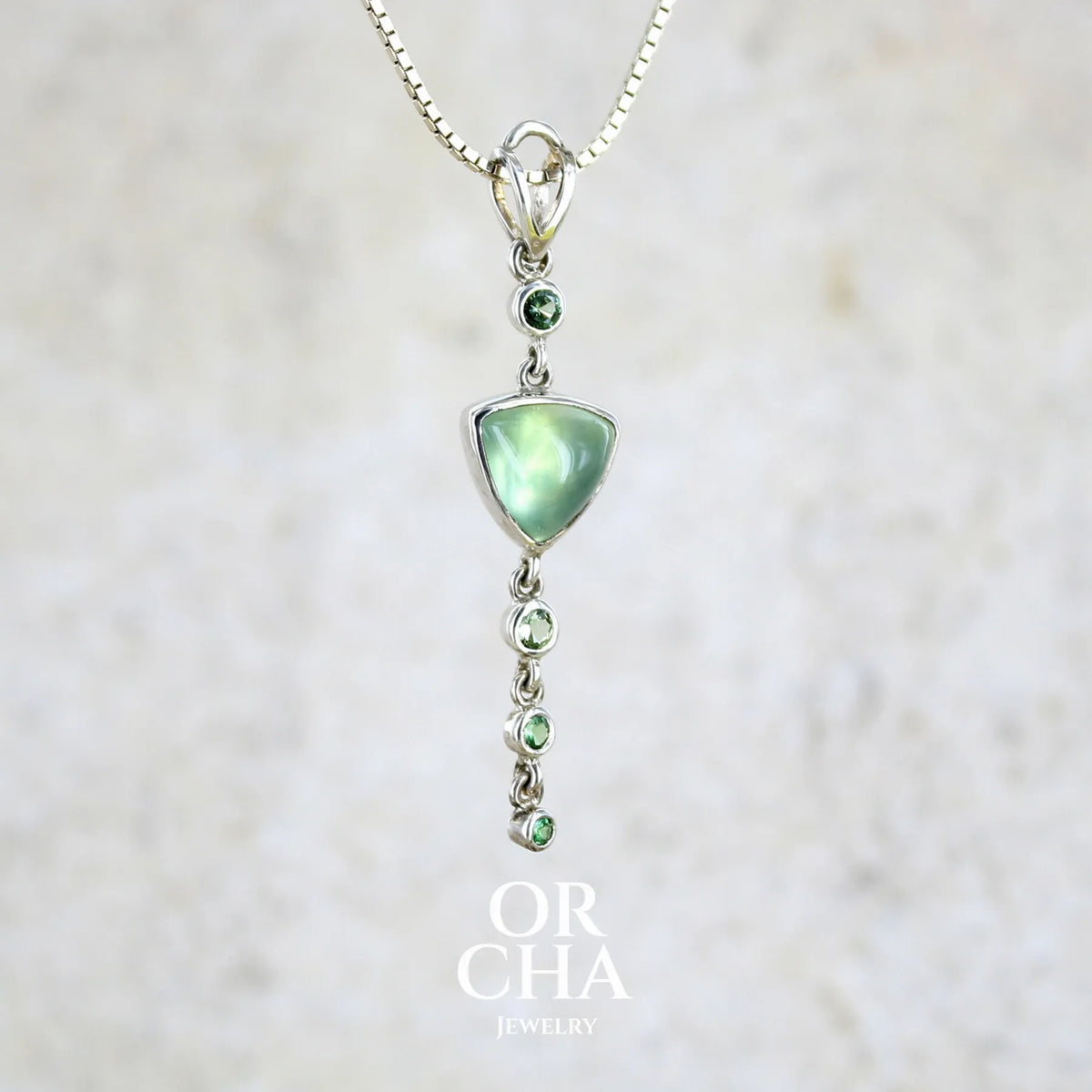Pendentif pour femme en argent massif sertie d'un cabochon triangulaire de Préhnite ainsi que de petites tourmalines vertes formant une cascade colorée vert pastel lumineux. Pierre transparente à inclusions. Bijou unique réalisé par Orchajewelry