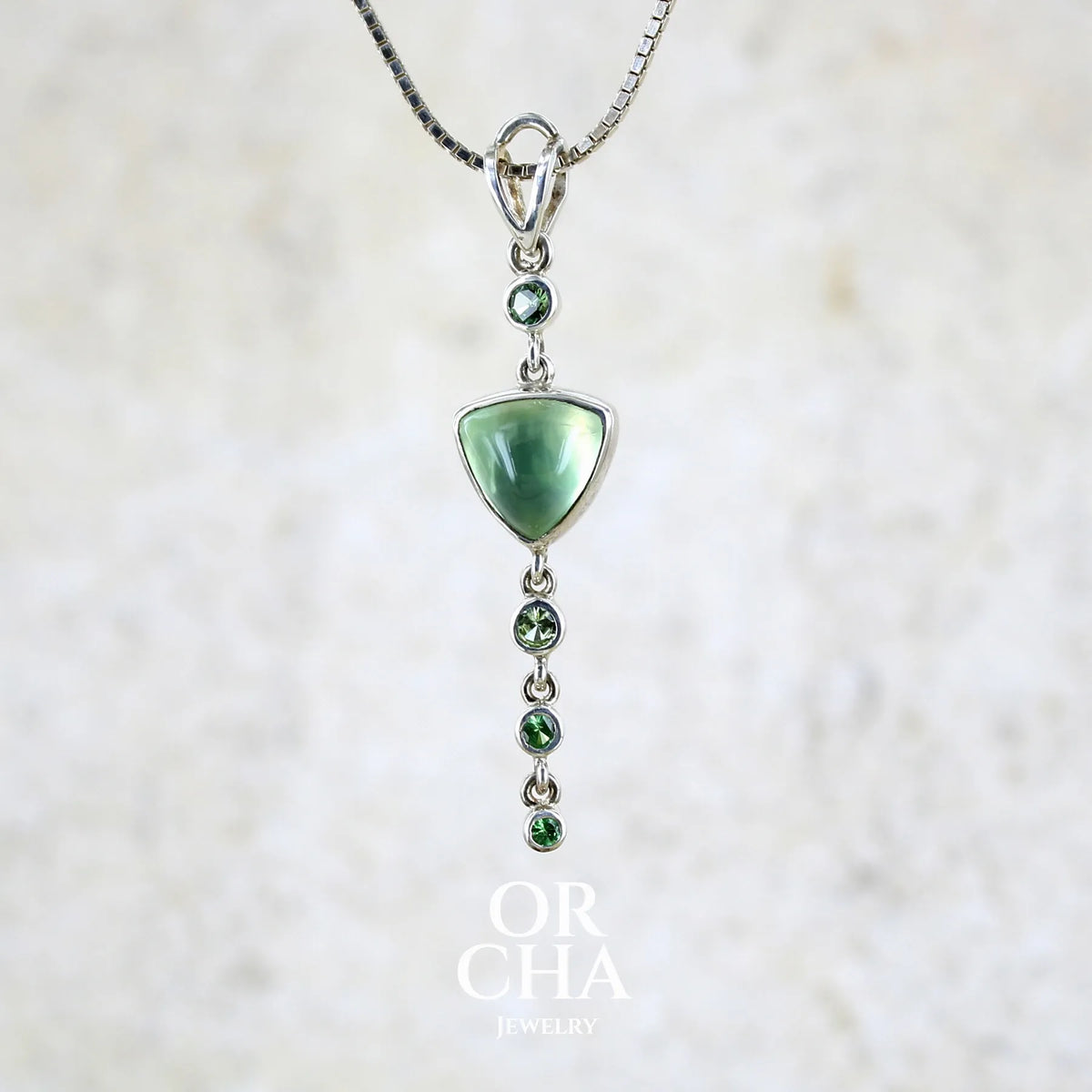 Pendentif pour femme en argent massif sertie d'un cabochon triangulaire de Préhnite ainsi que de petites tourmalines vertes formant une cascade colorée vert pastel lumineux. Pierre transparente à inclusions. Bijou unique réalisé par Orchajewelry