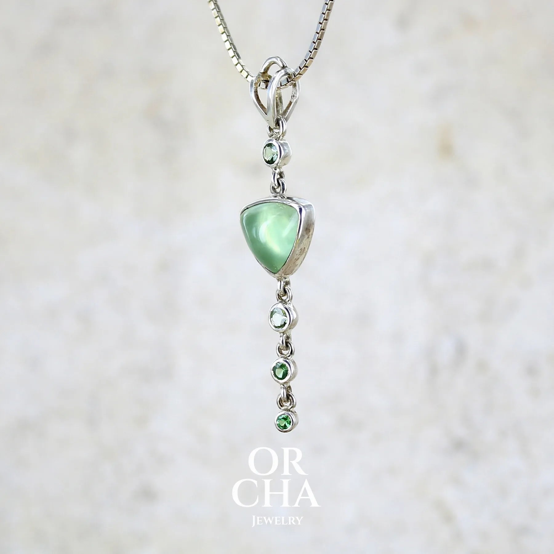 Pendentif pour femme en argent massif sertie d'un cabochon triangulaire de Préhnite ainsi que de petites tourmalines vertes formant une cascade colorée vert pastel lumineux. Pierre transparente à inclusions. Bijou unique réalisé par Orchajewelry