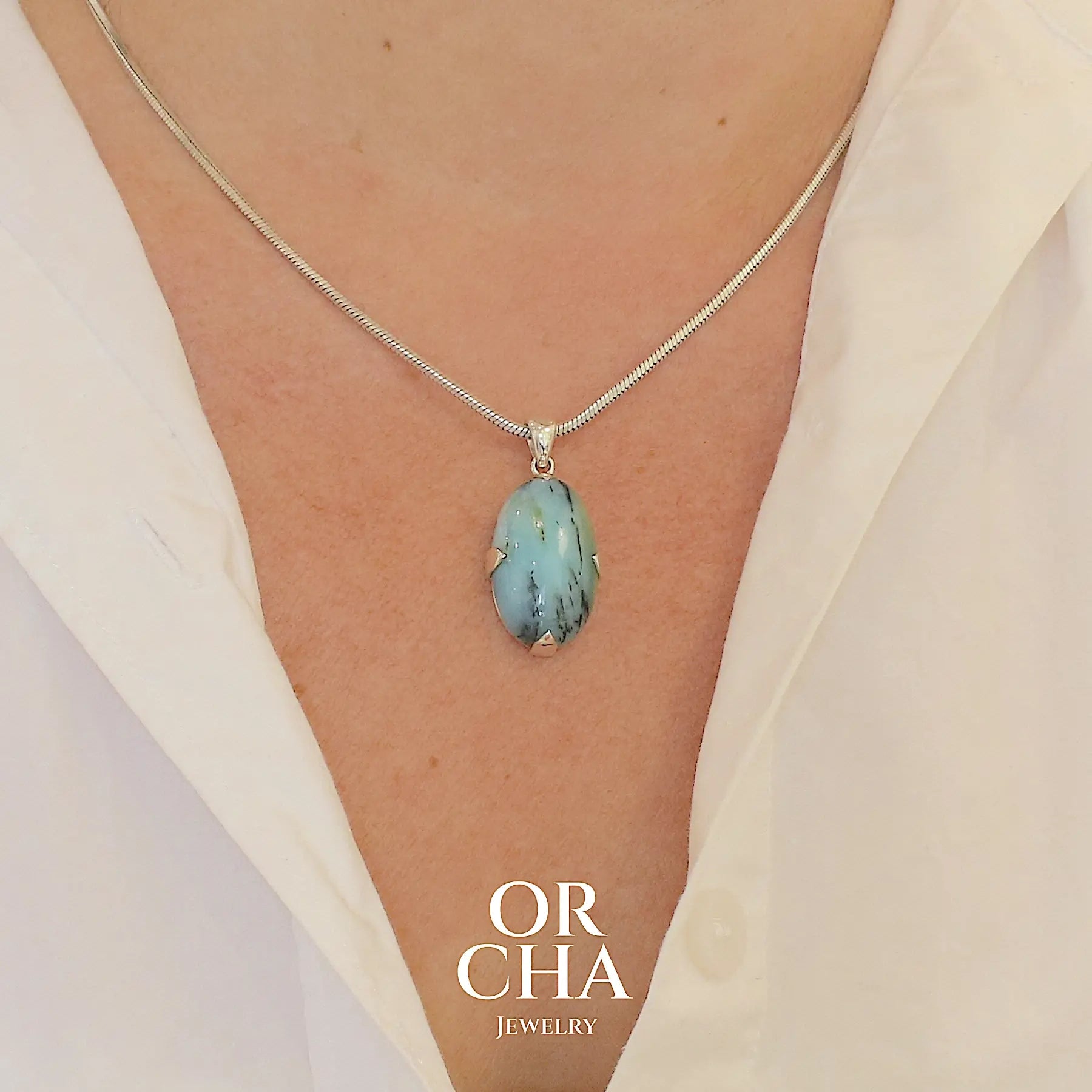 photo sur modèle d'un pendentif en argent massif, sertie d'un cabochon ovale d'opale bleue du Pérou de couleur bleu laiteux à inclusions de dendrites noires. Pierre opaque et translucide. Bijoux unique, éthique et durable présenté dans un coffret de notre marque, prêt à offrir. Livraison offerte, certificat d'authenticité, service client.