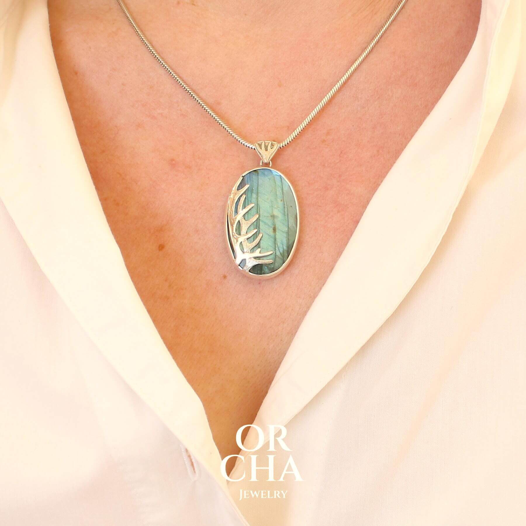 Pendentif sur mannequin, pour femme, en argent massif sertie d'une labradorite aux reflets bleus, Une ramure de cerf sculptée en argent penchasse la pierre. Pierre facettée naturelle translucide présentant des inclusions fréquents sur ce type de pierre. Bijoux unique, éthique et durable présenté dans un coffret de notre marque, prêt à offrir. Livraison offerte, certificat d'authenticité, service client.
