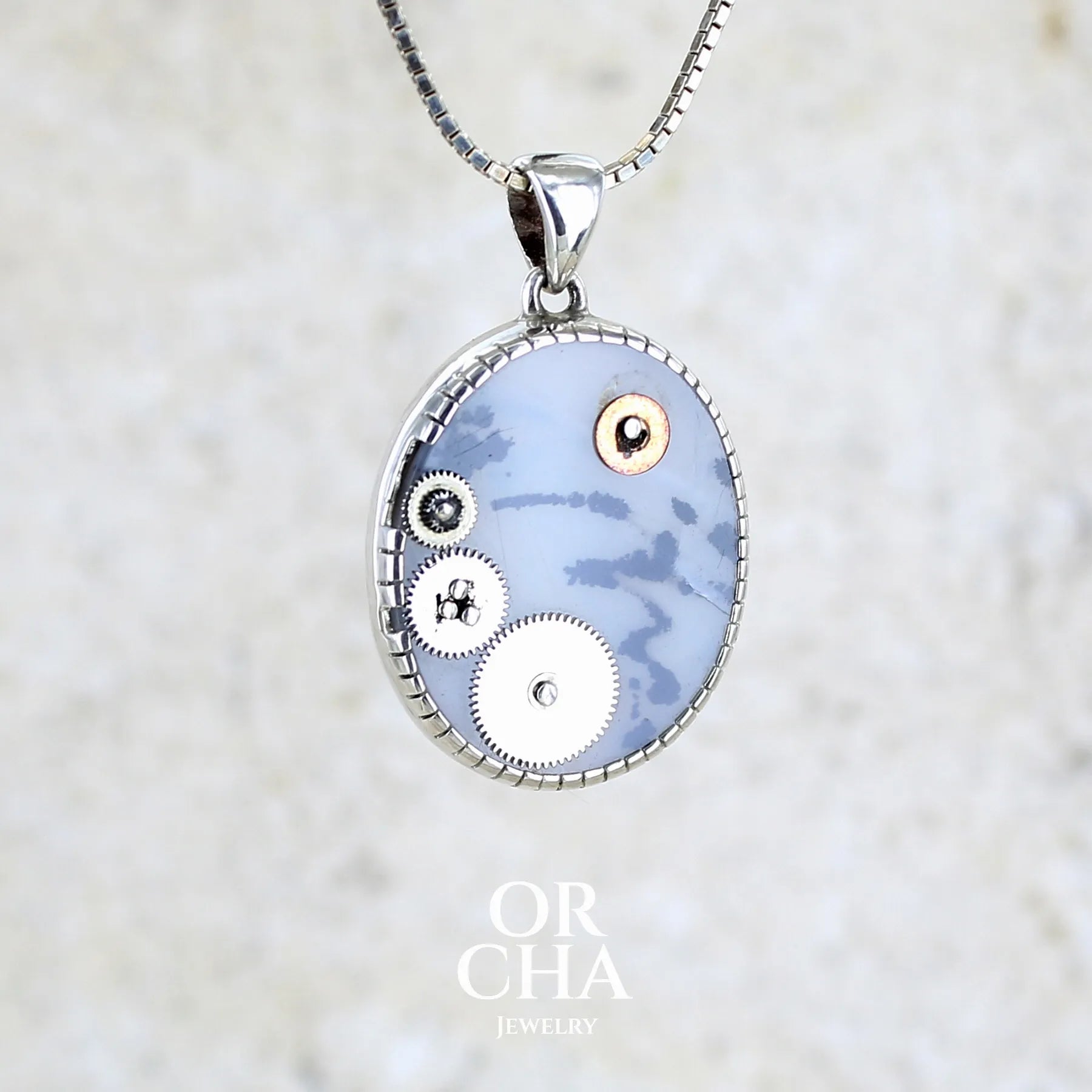 Pendentif steam punk pour homme en argent massif sertie d'une tranche ovale d'agate dendritique de couleur gris et noir, orné de mécanismes de montre. Pierre translucide à inclusions de type tâches d'encre de dendrites de manganèse noires contrastées. Bijou unique réalisé par Orchajewelry