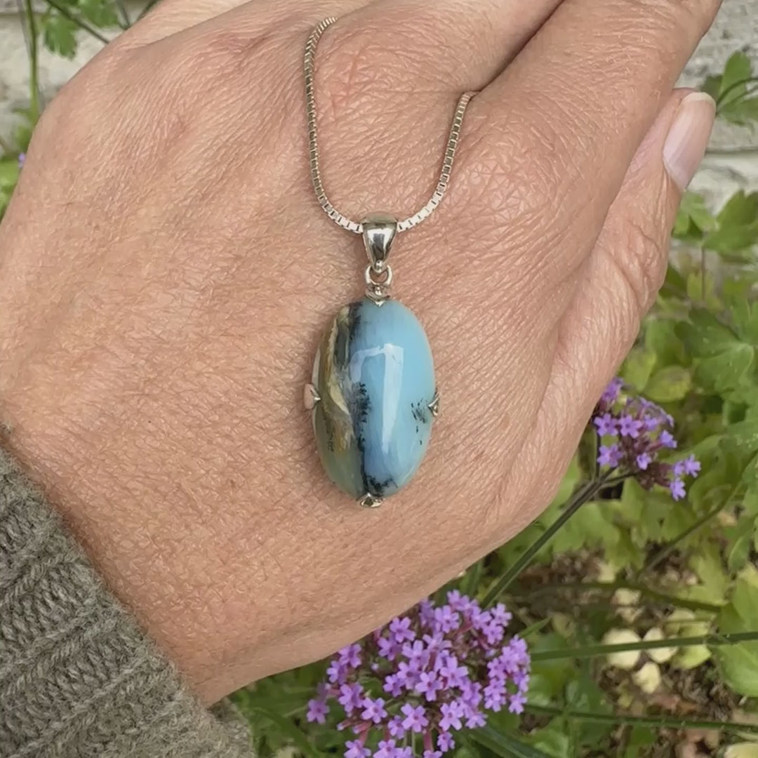 vidéo d'un pendentif pour femme et homme en argent massif, sertie d'un cabochon ovale d'opale bleue du Pérou de couleur bleu laiteux à inclusions de dendrites paysages noires et de roche mère marron. Pierre opaque et translucide. Bijoux unique, éthique et durable présenté dans un coffret de notre marque, prêt à offrir. Livraison offerte, certificat d'authenticité, service client.
