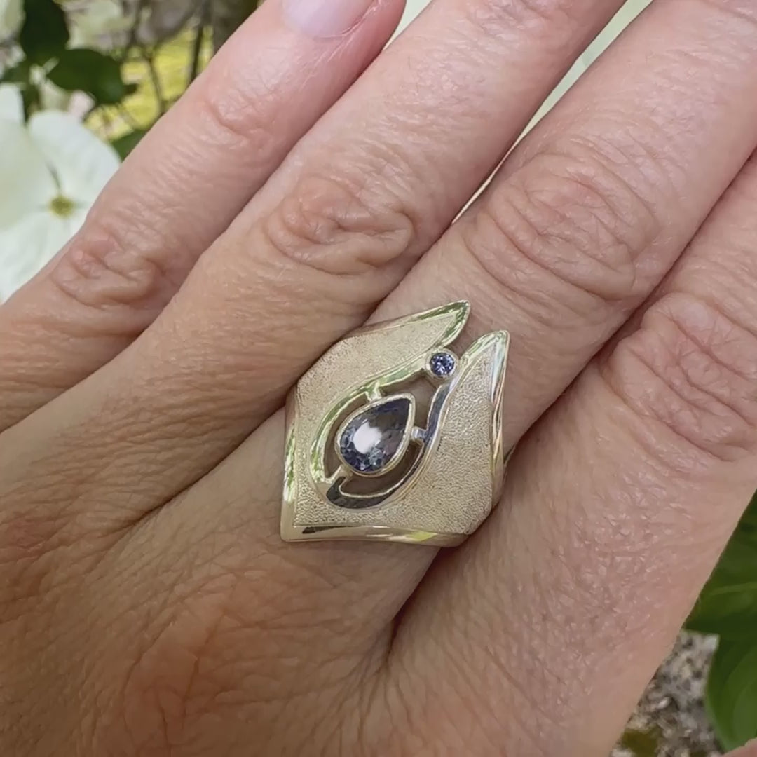 Vidéo d'une grande bague pour femme, fantaisy luxe en argent massif, sertie de tanzanites bleu lavande. Pierre facettée en forme de goutte et une plus petite ronde. Pierre  lumineuse et propre à l'oeil. Taille 59. Bijoux unique, éthique et durable présenté dans un coffret de notre marque, prêt à offrir. Livraison offerte, certificat d'authenticité, service client.