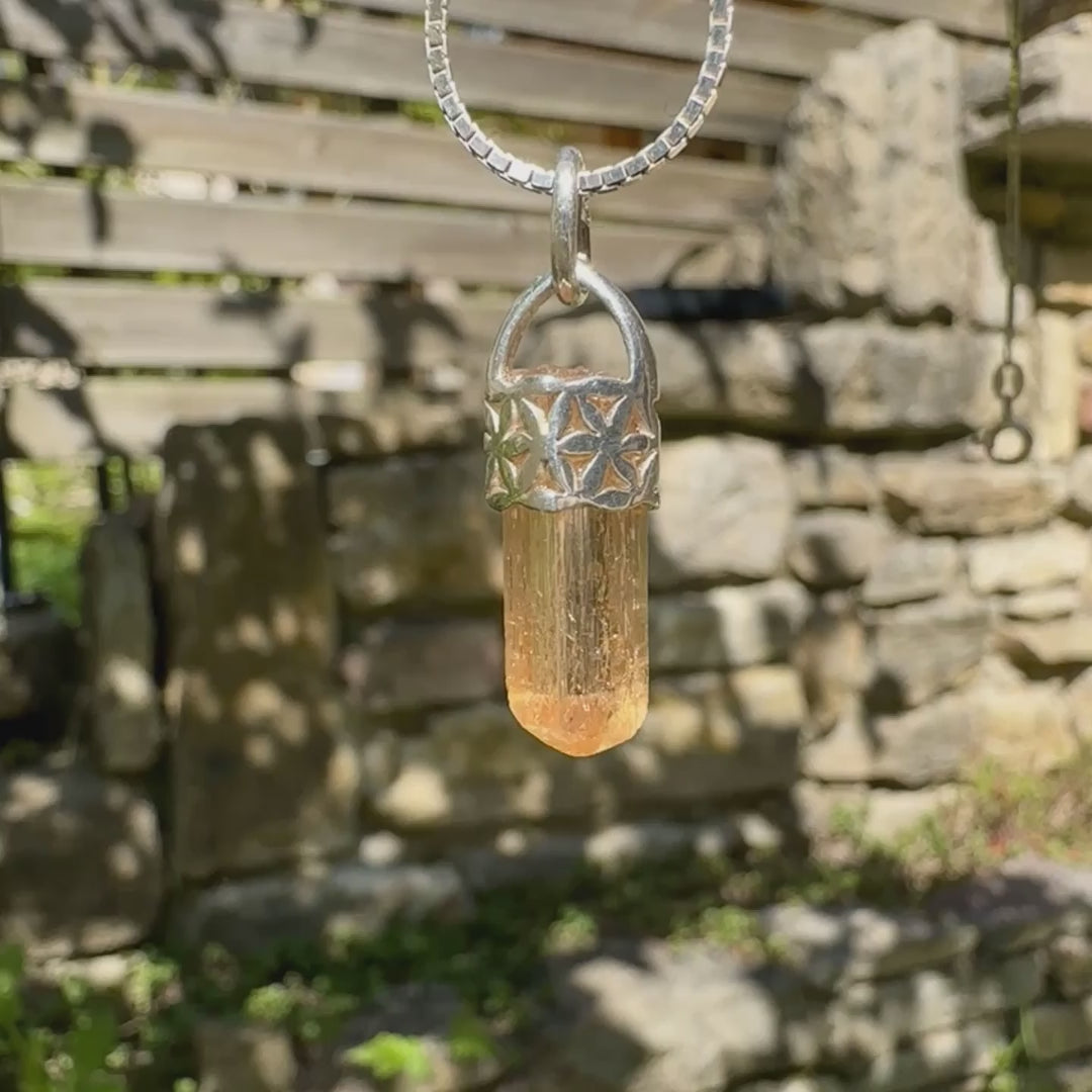 Vidéo d'un pendentif cristal de topaze impériale en argent massif, sertissage filigrané de fleurs de vie. La pierre est  de couleur jaune arrangé pêche. Cristal brute, transparent, inclusions naturelles. Bijoux unique, éthique et durable présenté dans un coffret de notre marque, prêt à offrir. Livraison offerte, certificat d'authenticité, service client.