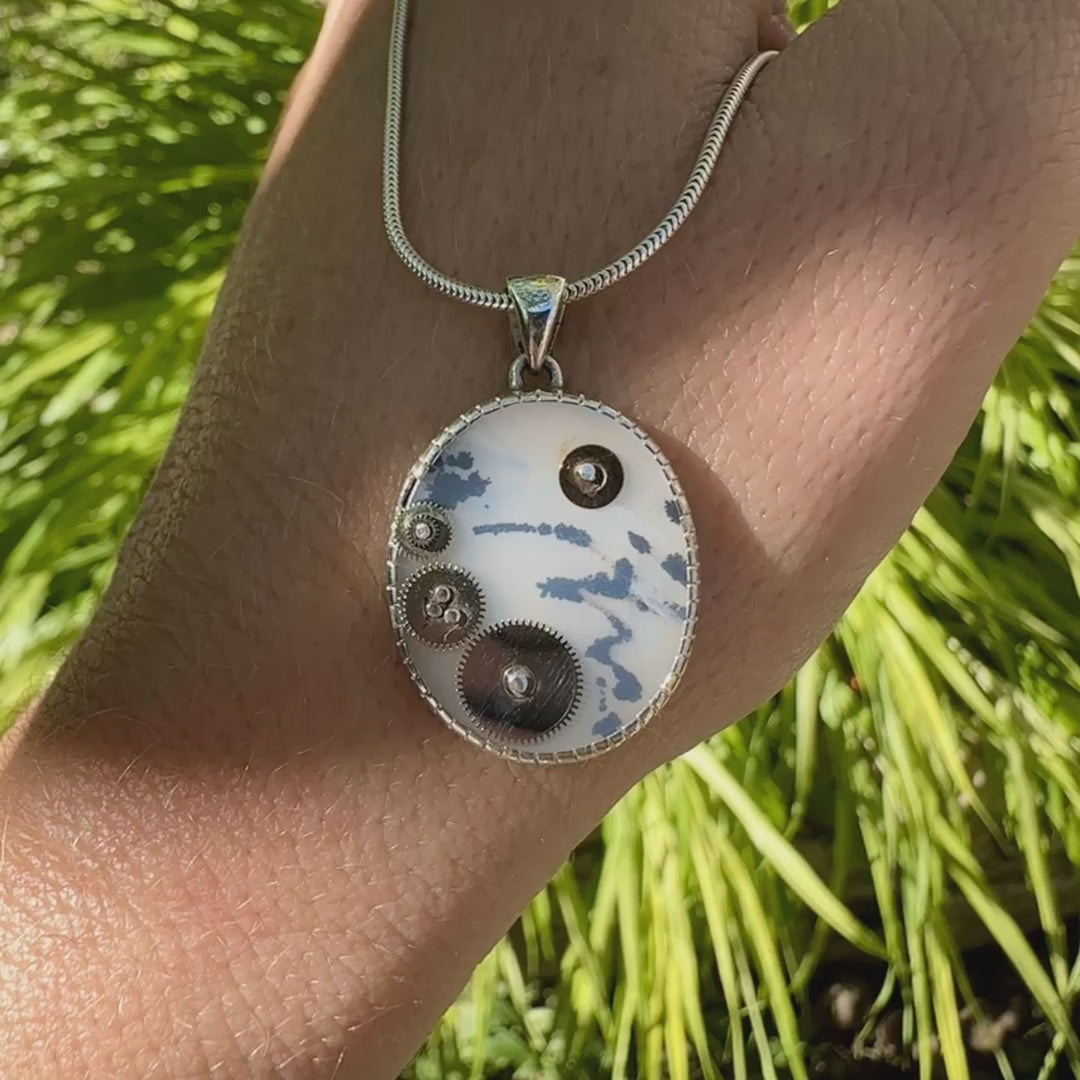 Pendentif steam punk pour homme en argent massif sertie d'une tranche ovale d'agate dendritique de couleur gris et noir, orné de mécanismes de montre. Pierre translucide à inclusions de type tâches d'encre de dendrites de manganèse noires contrastées. Bijou unique réalisé par Orchajewelry