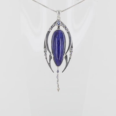 Pendentif contemporain en argent massif représentant une forme organique stylisée. Il est orné d’une azurite bleu profond de forme ovale allongée, encadrée de deux structures symétriques serties de petites tanzanite rondes violettes. Une ligne de pierres descend sous la pierre centrale, se terminant par un élément pointu sculpté. Bijoux unique, éthique et durable présenté dans un coffret de notre marque, prêt à offrir. Livraison offerte, certificat d'authenticité, service client.