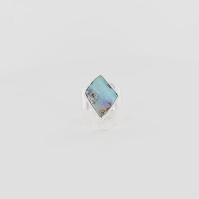 Bague en argent massif 925 sertie d'une opale boulder australienne de forme diamant asymétrique. La pierre présente une magnifique iridescence bleu-vert dominante avec des reflets violacés et quelques inclusions dorées naturelles. La monture simple et élégante en argent poli encadre la pierre, mettant en valeur ses couleurs changeantes. Bague réalisée en taille 56. Bijoux unique, éthique et durable présenté dans un coffret de notre marque, prêt à offrir. Livraison offerte, certificat d'authenticité, service