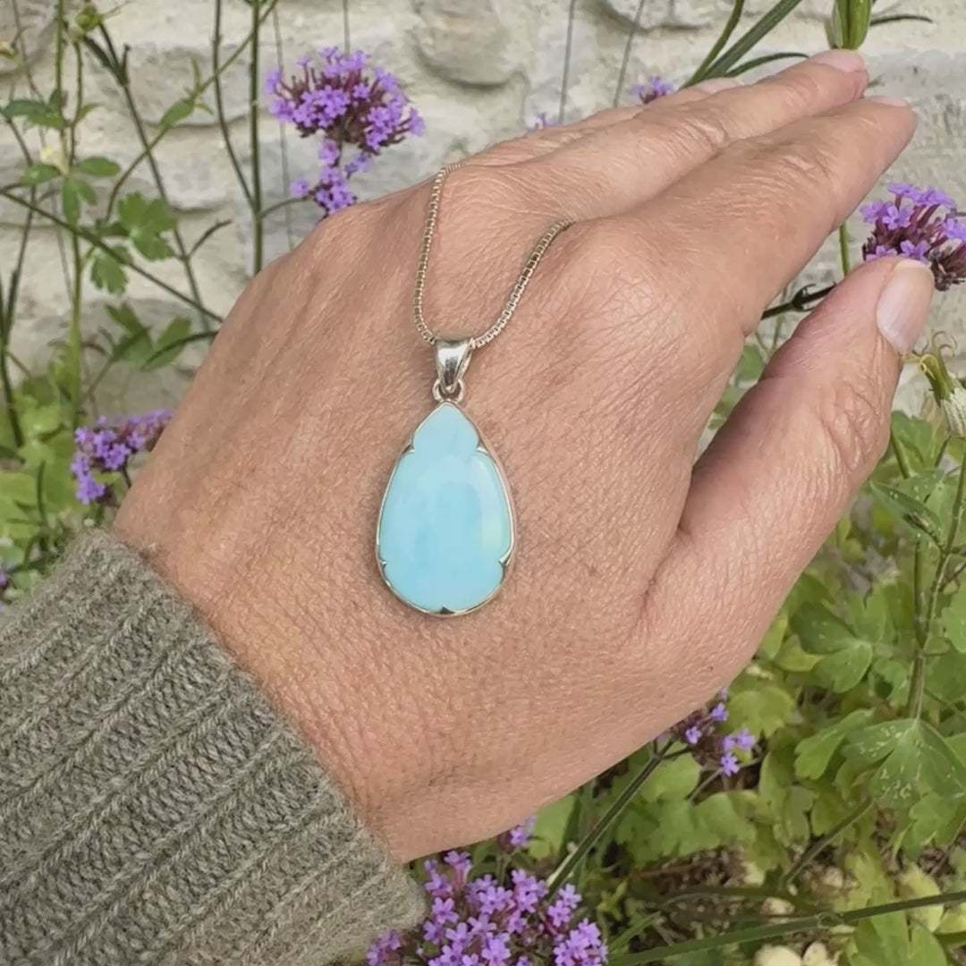 photo sur modèle d'un pendentif en argent massif, sertie d'un cabochon d'opale bleue du Pérou de couleur bleu ciel clair. Pierre en forme de goutte. Pierre opaque et translucide. Bijoux unique, éthique et durable présenté dans un coffret de notre marque, prêt à offrir. Livraison offerte, certificat d'authenticité, service client.