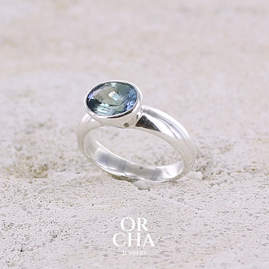 profil gauche d'une bague solitaire pour femme en argent massif sertie d'une tanzanite bicolore. Pierre facettée de forme ovale de qualité transparente, propre à l'oeil. Pierre naturelle de couleur bleu verte et logés reflets violets. Bague réalisée en taille 50. Bijoux unique, éthique et durable présenté dans un coffret de notre marque, prêt à offrir. Livraison offerte, certificat d'authenticité, service client.