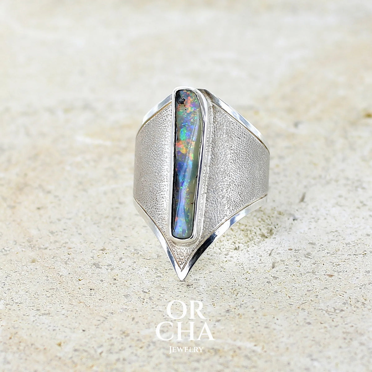 Profil gauche d'une bague plastron pour femme en argent massif 925 sertie d'une opale boulder aux feux multicolores. Cabochon d'opale massive naturelle, de qualité opaque. Bijoux unique réalisé en taille 59 par Orcha Jewelry.