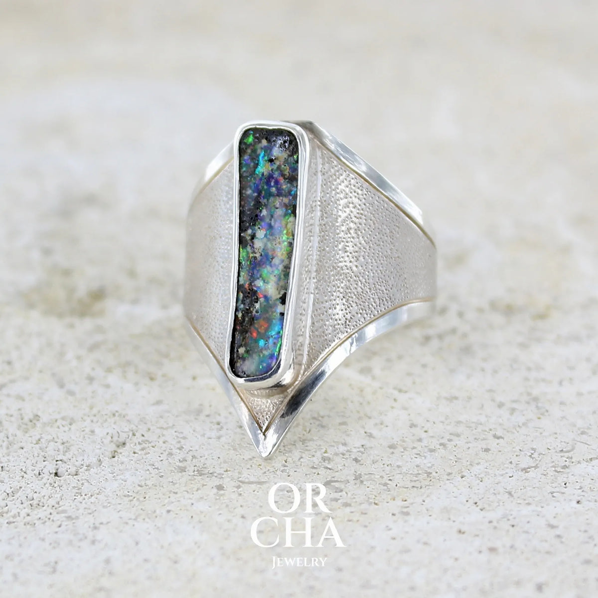 Profil gauche d'une bague pour femme en argent massif 925 sertie d'une opale boulder aux feux multicolores. Cabochon d'opale massive naturelle taillée en forme de rectangle.. Pierre de qualité opaque. Bague unique réalisée en taille 55 par Orcha Jewelry.