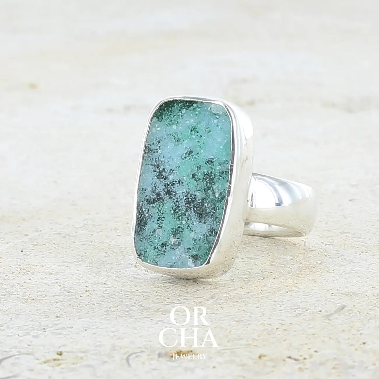 Profil gauche d'une bague pour femme en argent massif sertie d'un magnifique gemsilica druzy: chrysocale solidifiée recouverte de fins cristaux de quartz scintillant. Pierre de qualité opaque taillée en tranche de forme rectangulaire. Pierre naturelle originaire de République dominicaine du Congo de couleur turquoise, verte et noire. Taille 56. Bijoux unique, éthique et durable présenté dans un coffret de notre marque, prêt à offrir. Livraison offerte, certificat d'authenticité, service client.