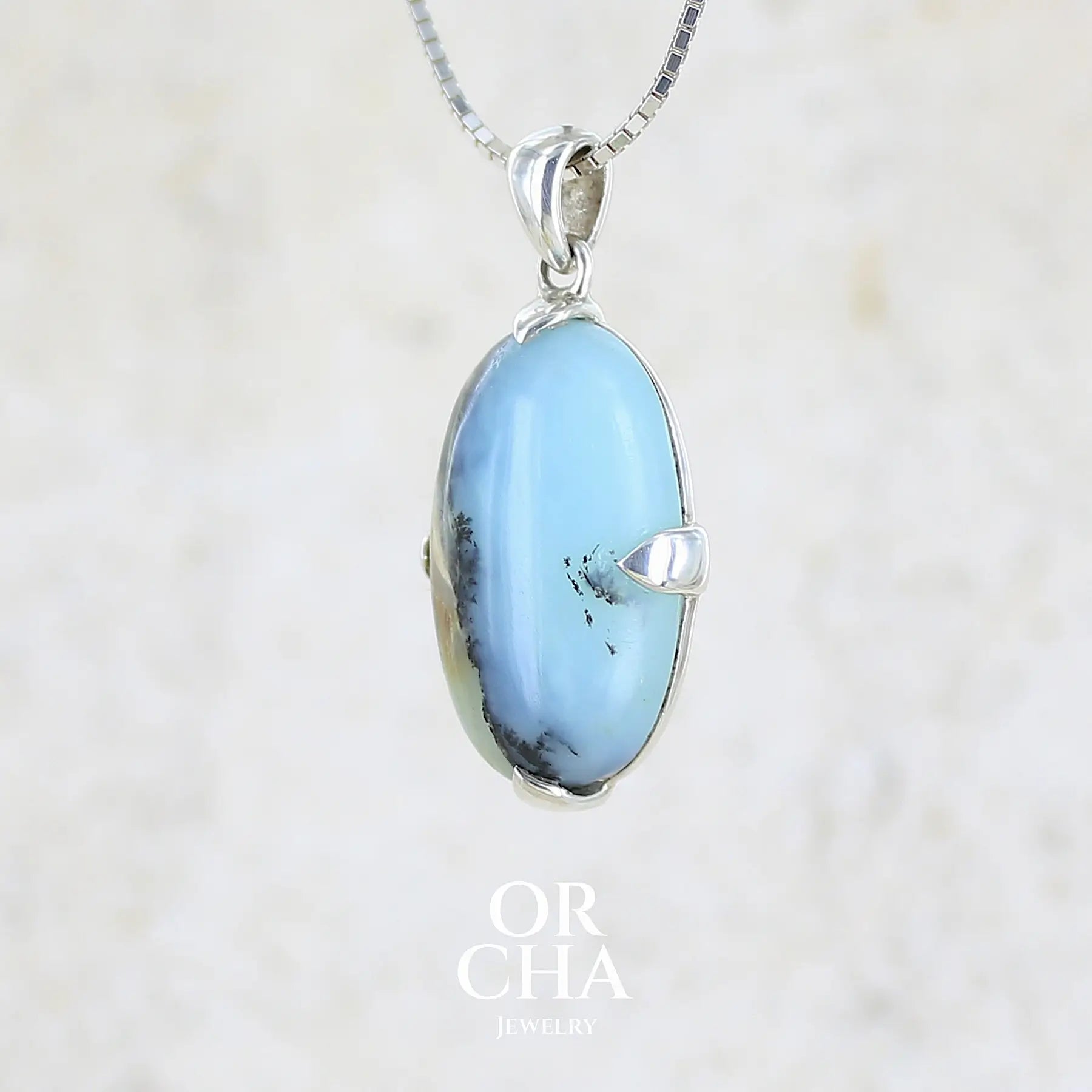 profil gauche d'un pendentif pour femme et homme en argent massif, sertie d'un cabochon ovale d'opale bleue du Pérou de couleur bleu laiteux à inclusions de dendrites paysages noires et de roche mère marron. Pierre opaque et translucide. Bijoux unique, éthique et durable présenté dans un coffret de notre marque, prêt à offrir. Livraison offerte, certificat d'authenticité, service client.