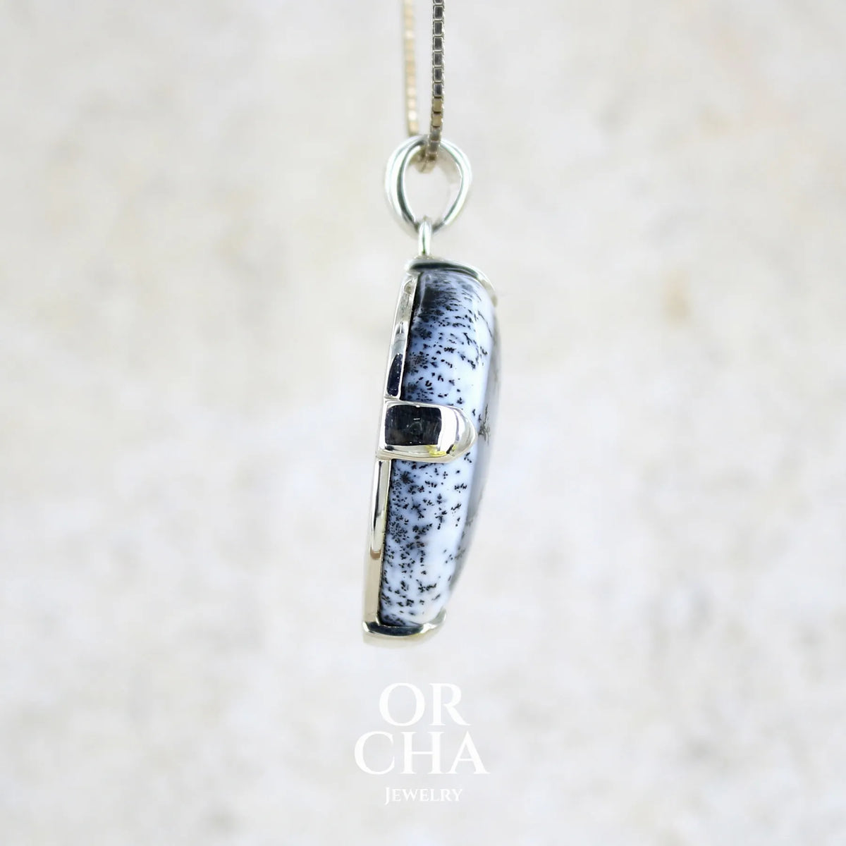 Profil d'un pendentif pour femme en argent massif sertie d'un cabochon en forme de goutte inversée d'agate dendritique de couleur blanc pur et noir. Pierre opaque à translucide à inclusions paysages de dendrites de manganèse noires contrastées. Bijou unique réalisée par Orchajewelry