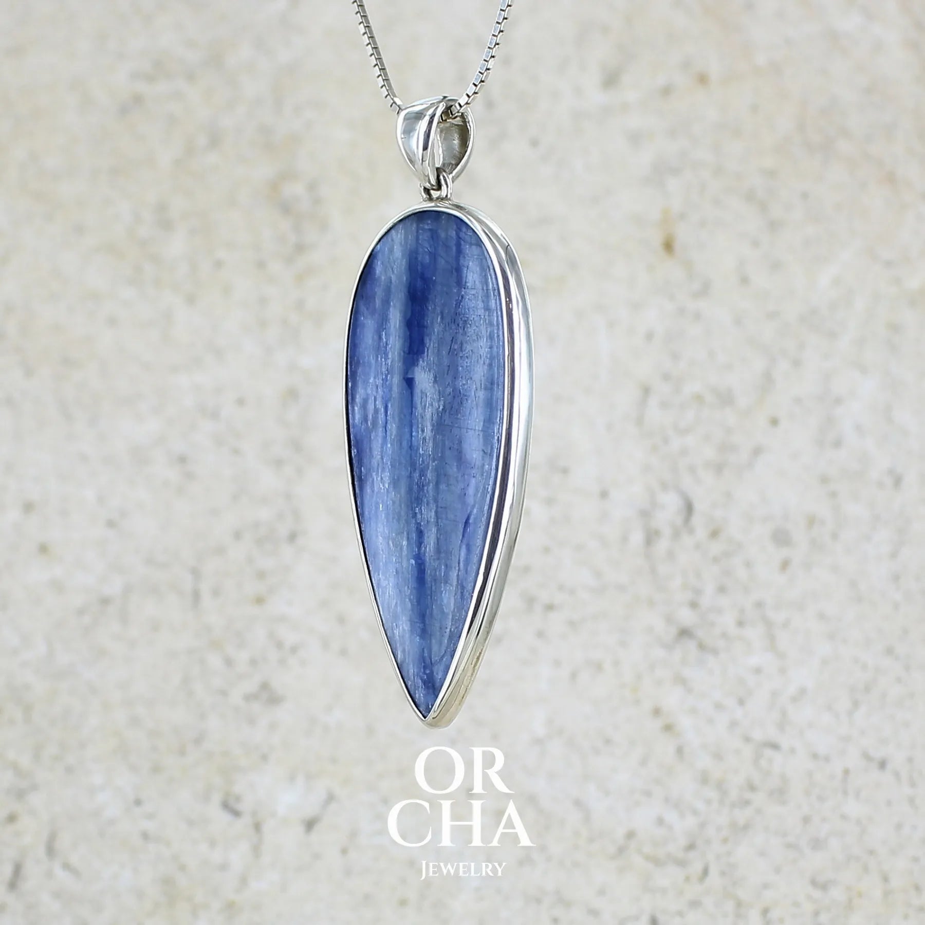 profil gauche d'un pendentif en argent massif serti d'un cabochon de cyanite de couleur bleue jeans nacré. Pierre de qualité translucide taillée en forme de goutte inversée comportant des inclusions. Bijoux unique par Orchajewelry
