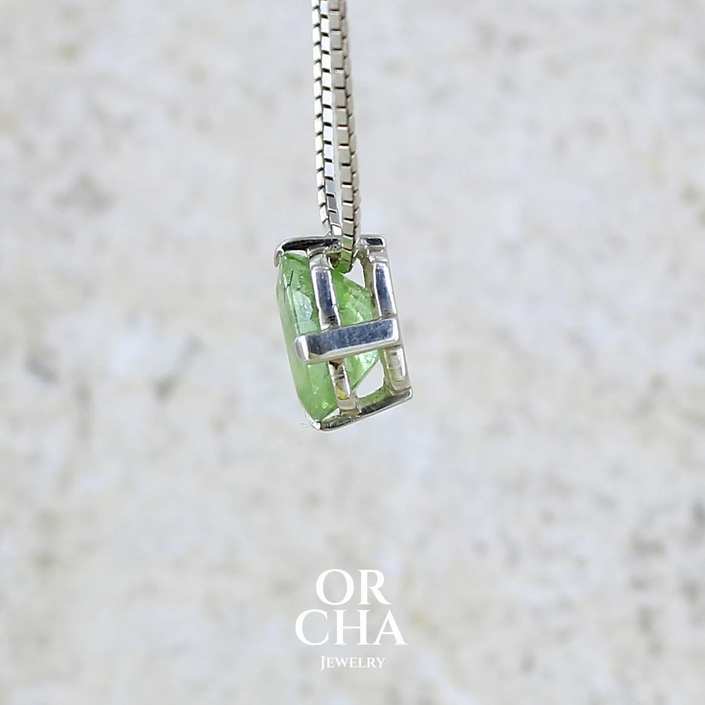 profil d'un collier solitaire pour femme en argent massif sertie d'un Péridot de couleur vert pastel anisé. Pierre facettée de forme carrée, qualité transparente présentant des inclusions. Pendentif monté sur une chaine en argent sterling de type maille miroir de 1,3 mm d'épaisseur et de 40cm de longueur. Bijoux unique réalisé par Orchajewelry