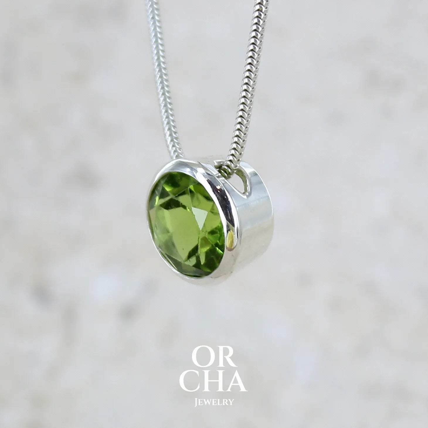 profil gauche d'un collier solitaire pour femme en argent massif serti d'un Péridot facettée de 4 carats. Pierre de forme ronde de qualité transparente, propre à l'oeil. Pierre naturelle de couleur vert olive doré intense et lumineuse. Pendentif monté sur une chaine en argent sterling de type maille serpent de 0,43 mm d'épaisseur et de 40 cm de longueur. Bijoux unique réalisé par Orchajewelry.
