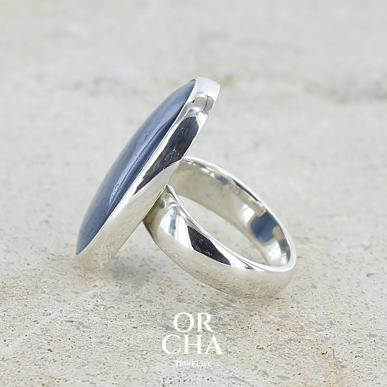 sertissage d'une bague pour femme en argent massif sertie d'un cabochon de cyanite de couleur bleu jean nacré. Pierre translucide à inclusions de forme ovale arrondie. Bijou unique réalisée en taille 58,5 par Orchajewelry