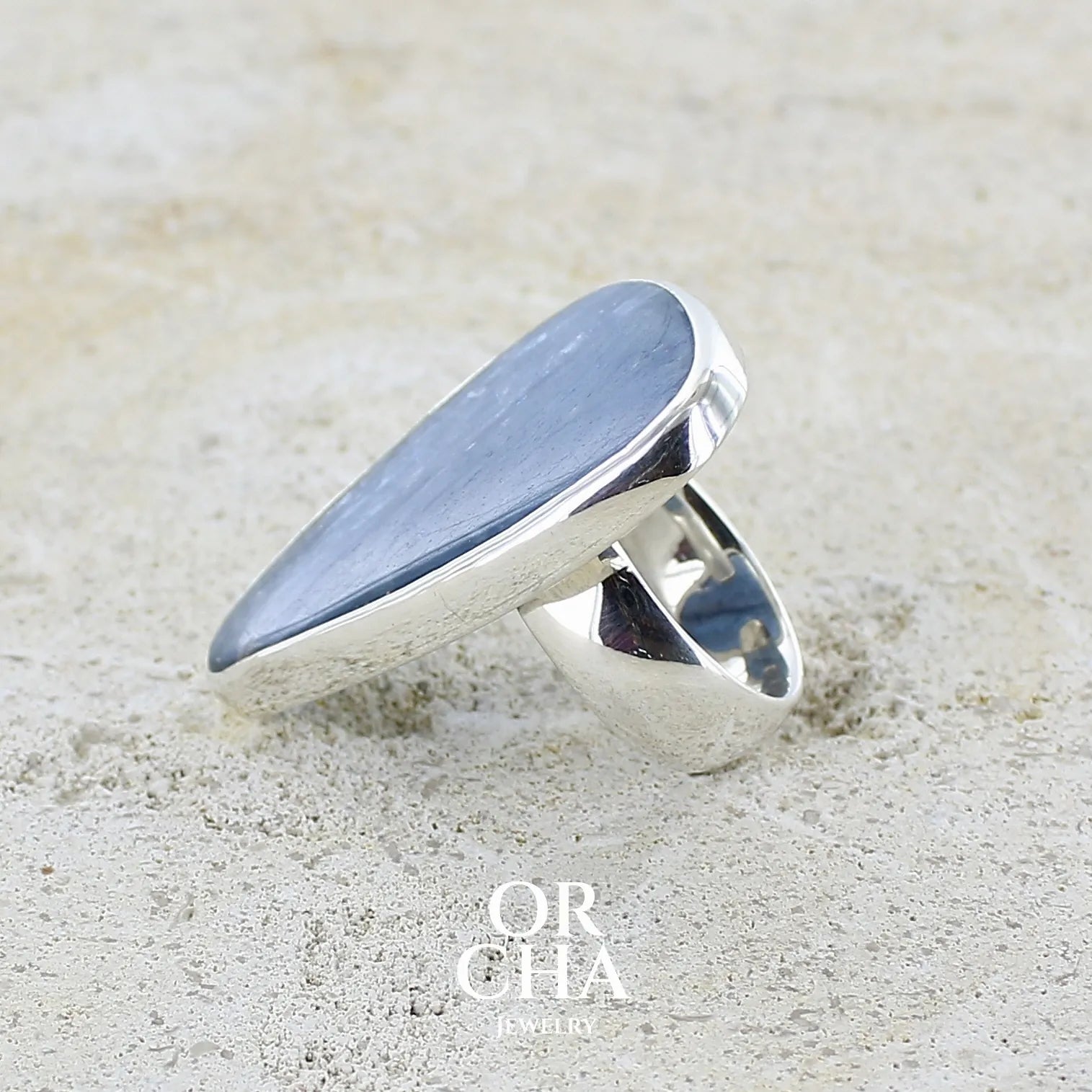 détail du sertissage d'une Bague longue et imposante pour femme en argent massif, sertie d'un cabochon de cyanite de couleur bleu jean nacré. Pierre taillée en forme de triangle de qualité translucide. Bijou unique réalisée en taille 58 par Orchajewelry