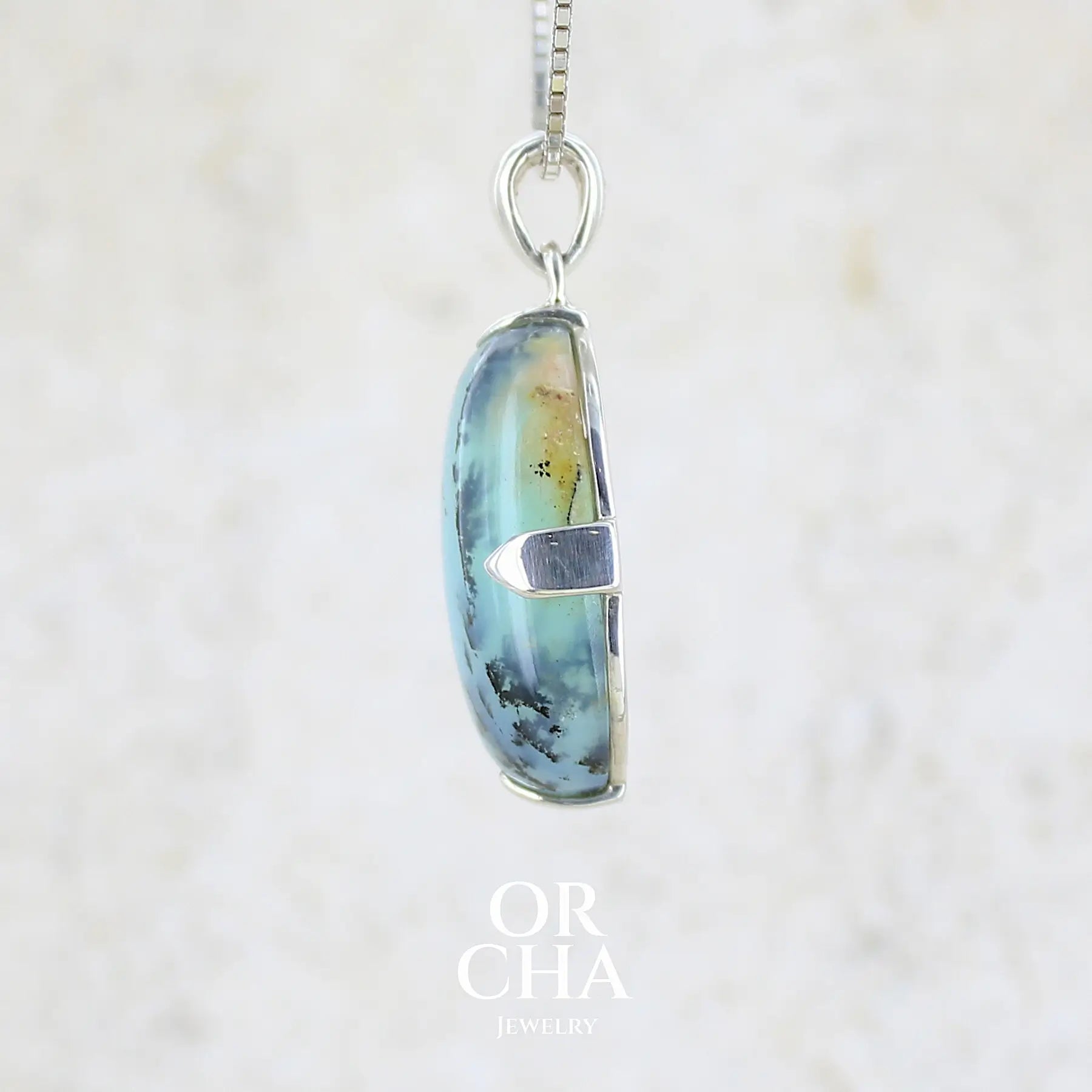 détail sertissage d'un pendentif en argent massif, sertie d'un cabochon ovale d'opale bleue du Pérou de couleur bleu laiteux à inclusions de dendrites noires. Pierre opaque et translucide. Bijoux unique, éthique et durable présenté dans un coffret de notre marque, prêt à offrir. Livraison offerte, certificat d'authenticité, service client.
